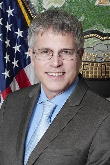 Portrait of a man in a gray suit, light blue shirt, and glasses, smiling. Background includes an American flag and a green and gold decorative wall.