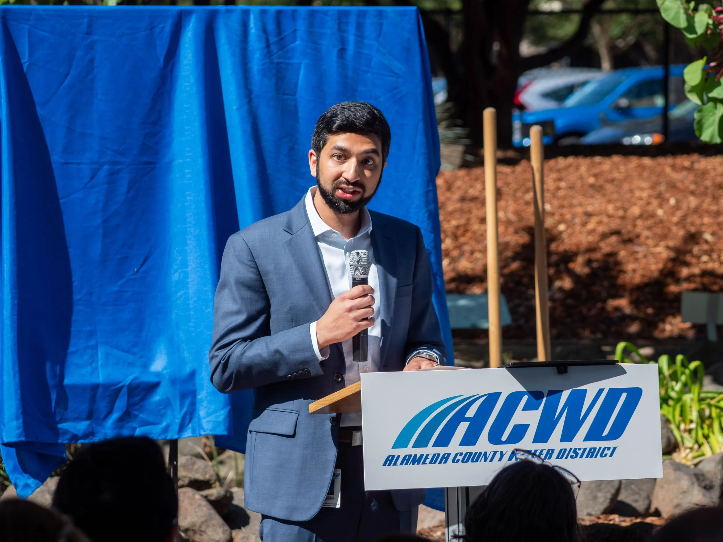 A man in a gray suit and white shirt speaking into a microphone at an outdoor event, standing behind a sign that reads "Alameda County Water District." There is a blue curtain or backdrop behind him, with parked cars and trees visible in the background.