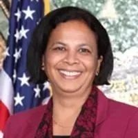 A woman smiling in front of an American flag, outdoors with trees in the background.