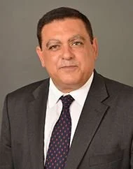 Professional headshot of a man in a dark suit, white shirt, and dotted tie against a gray background.