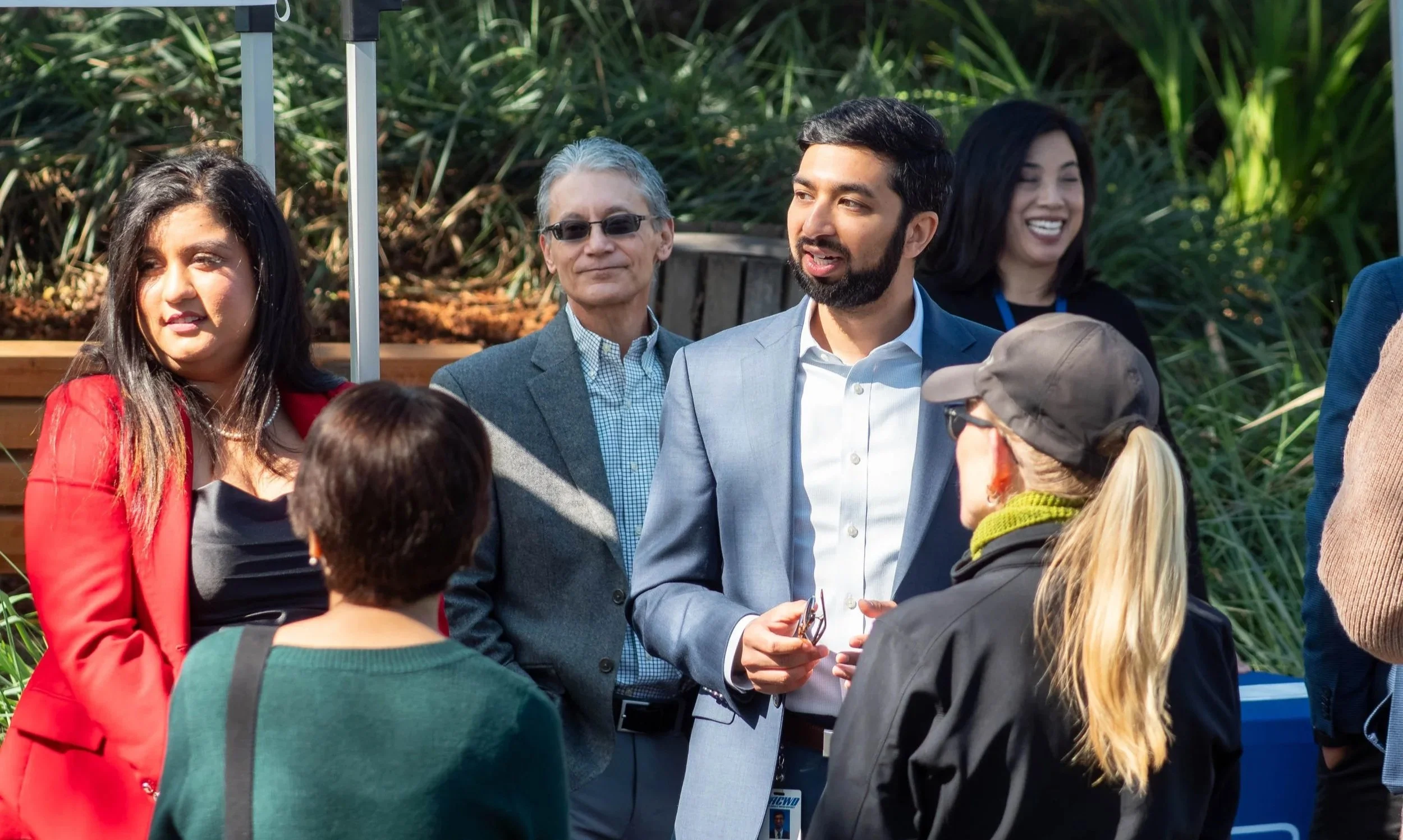 A group of diverse people engaging in conversation outdoors, with some wearing business attire, surrounded by green plants.