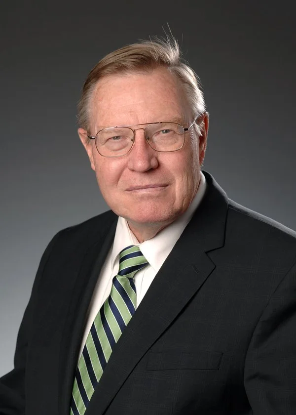 A portrait of an older man with light-colored hair, wearing glasses, a black suit, white shirt, and a green and navy blue striped tie, against a gray background.