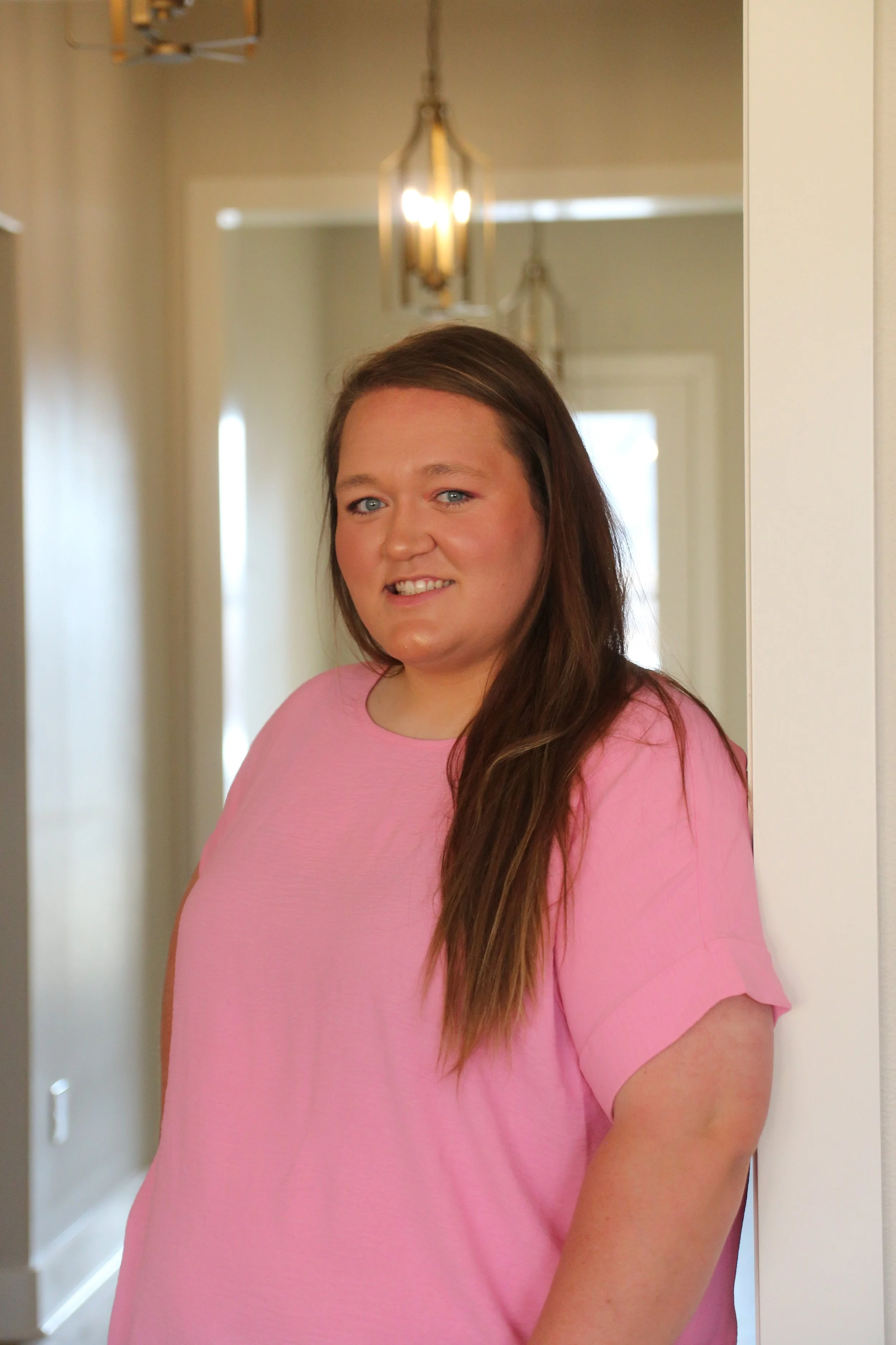 A woman with long brown hair and blue eyes wearing a pink top standing near a mirror in a brightly lit hallway.