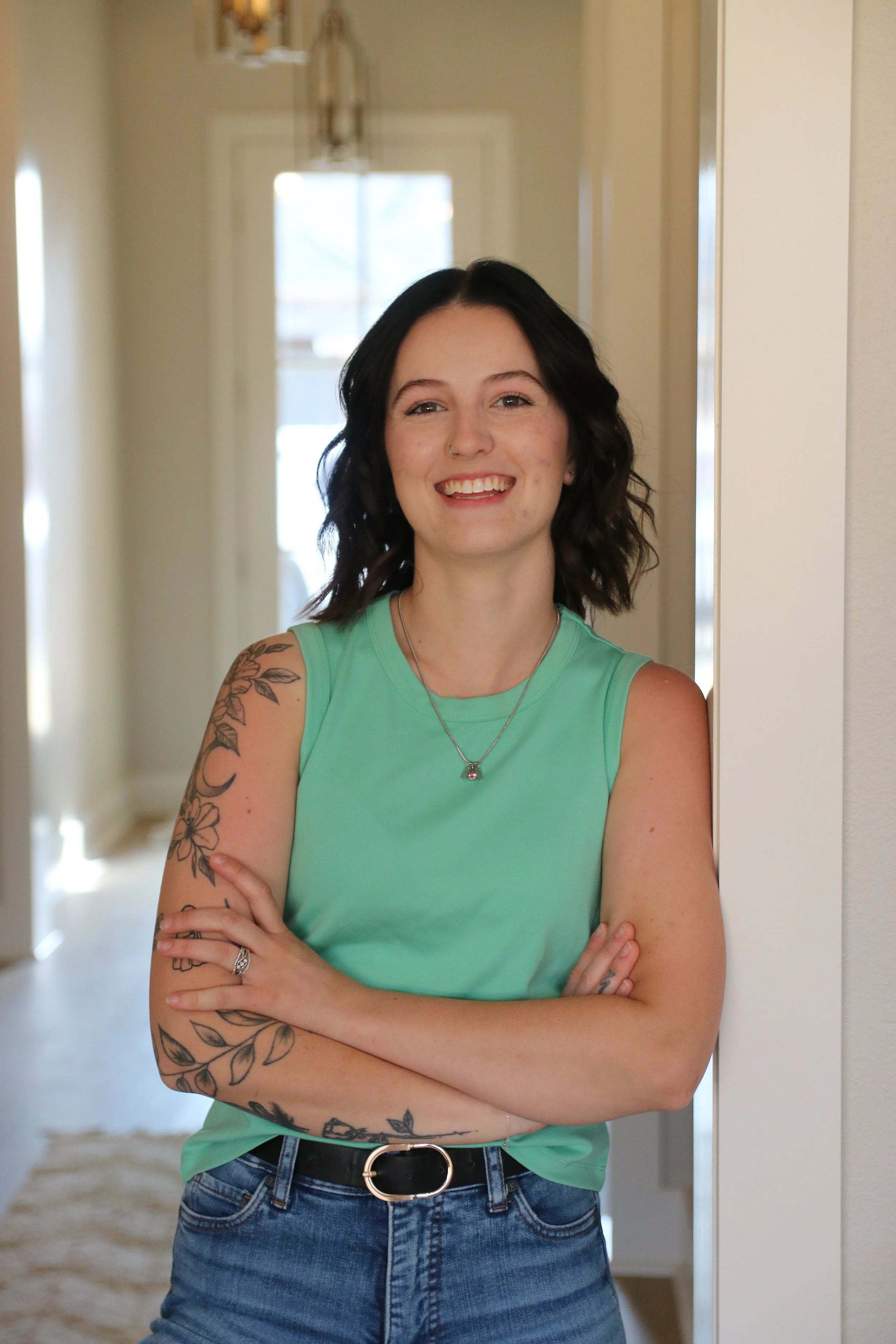 A woman with dark hair, tattoos on her arms, wearing a green sleeveless top, standing with crossed arms in a corridor with a window and a ceiling light in the background.