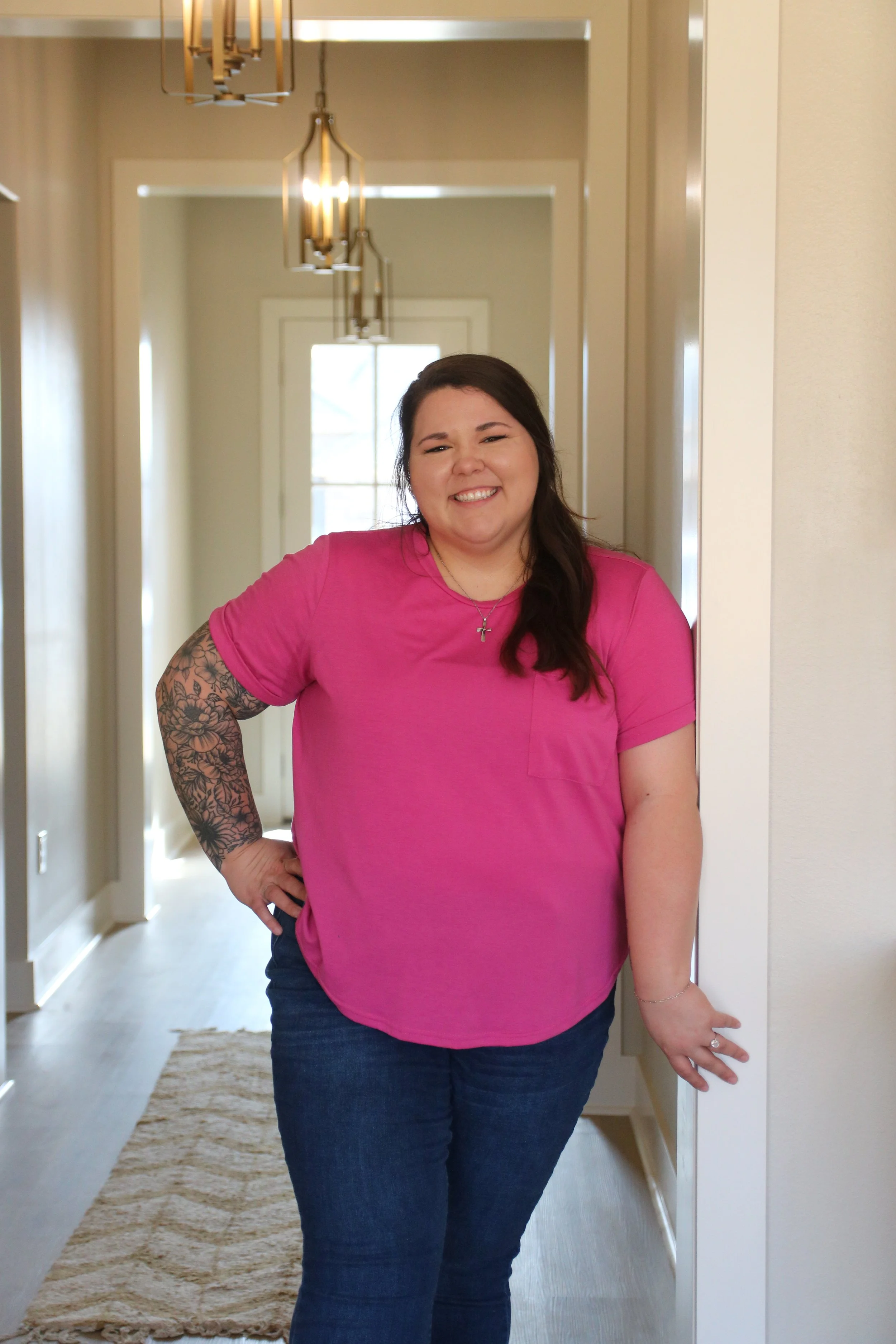 Smiling woman with tattooed arm wearing a pink T-shirt and jeans, standing in a hallway of a home, with pendant lights and a window in the background.