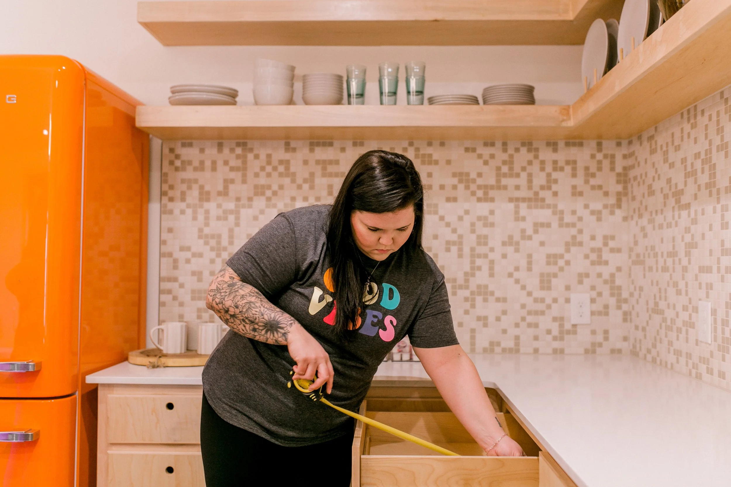 A woman with long dark hair, tattoos on her arm, wearing a gray T-shirt with colorful text, is working in a kitchen. She appears to be screwing or attaching something to a piece of furniture on a light wooden countertop. The kitchen has a modern design with open wooden shelves, a beige mosaic tile backsplash, and an orange refrigerator on the left side of the image.