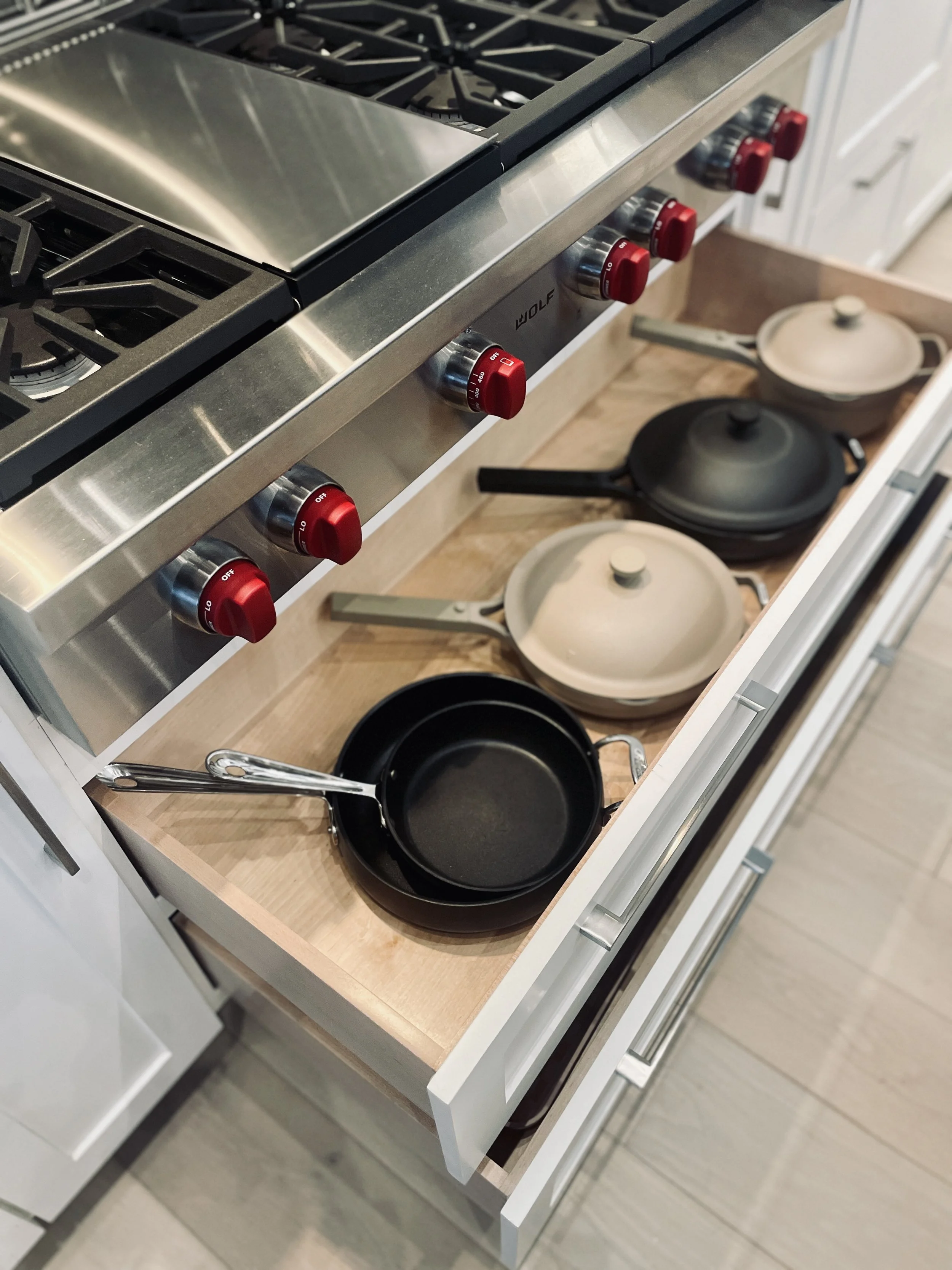 Stainless steel gas stove with four red knobs and black cast iron grates. Open drawer with black and beige pots and a black skillet on a wooden drawer bottom.