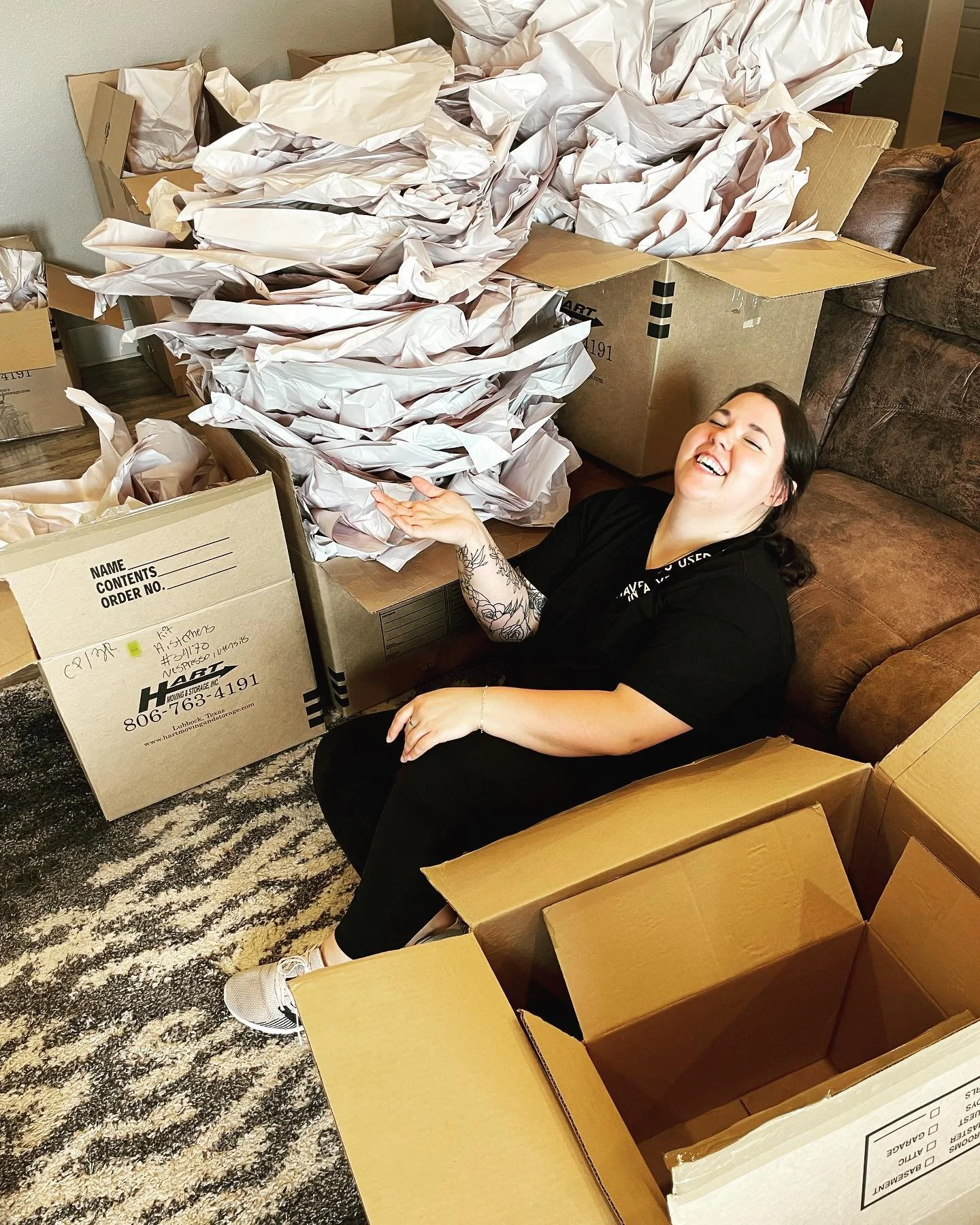 A woman sitting on the floor among open and partially filled cardboard boxes, with crumpled packing paper overflowing from some of the boxes, smiling and joyful.