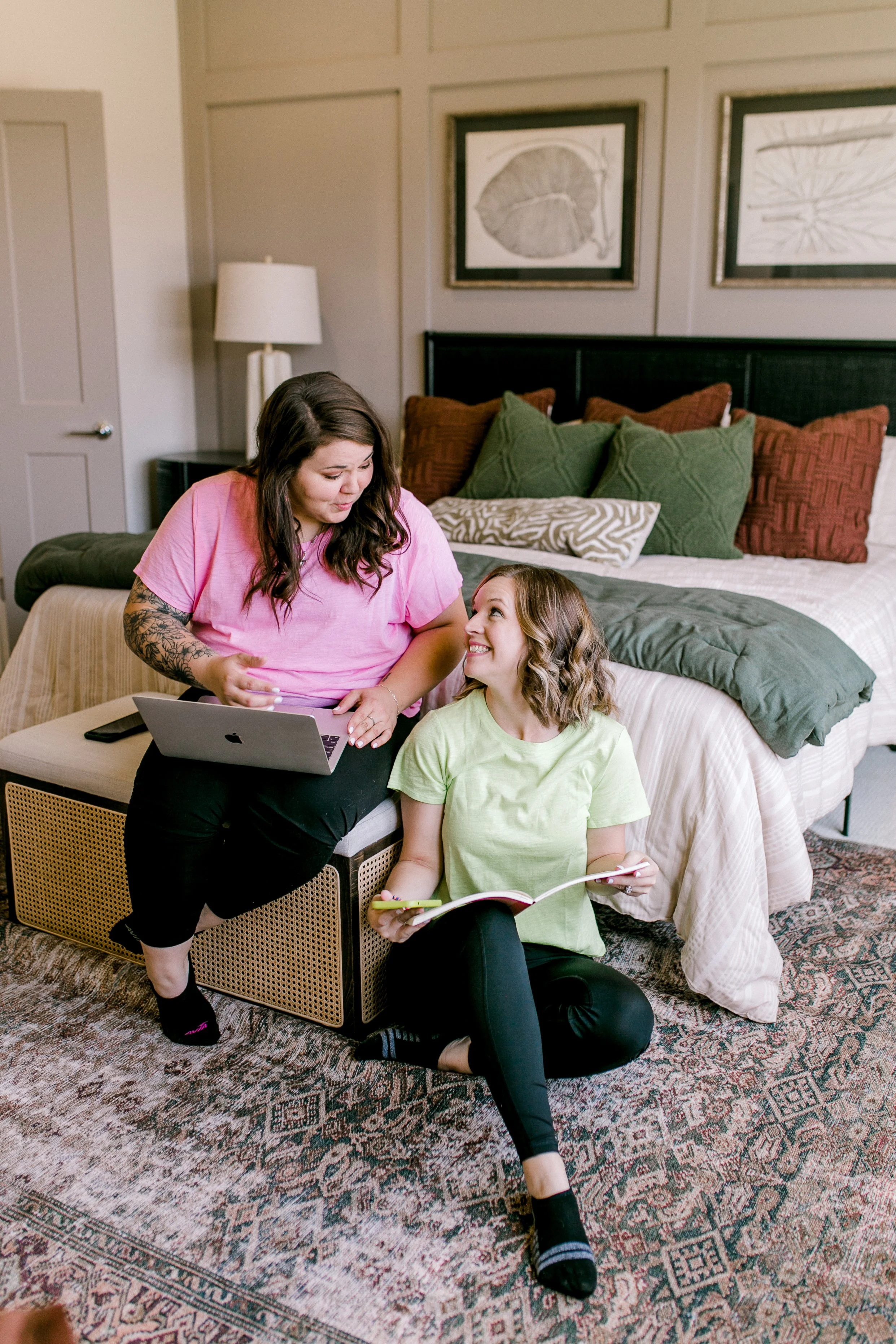 Two women and a young girl in a bedroom, with the women interacting with the girl. The women are using a laptop and a phone, while the girl holds a notebook or tablet. The bed in the background has multiple pillows and framed art on the wall, with a patterned rug on the floor.