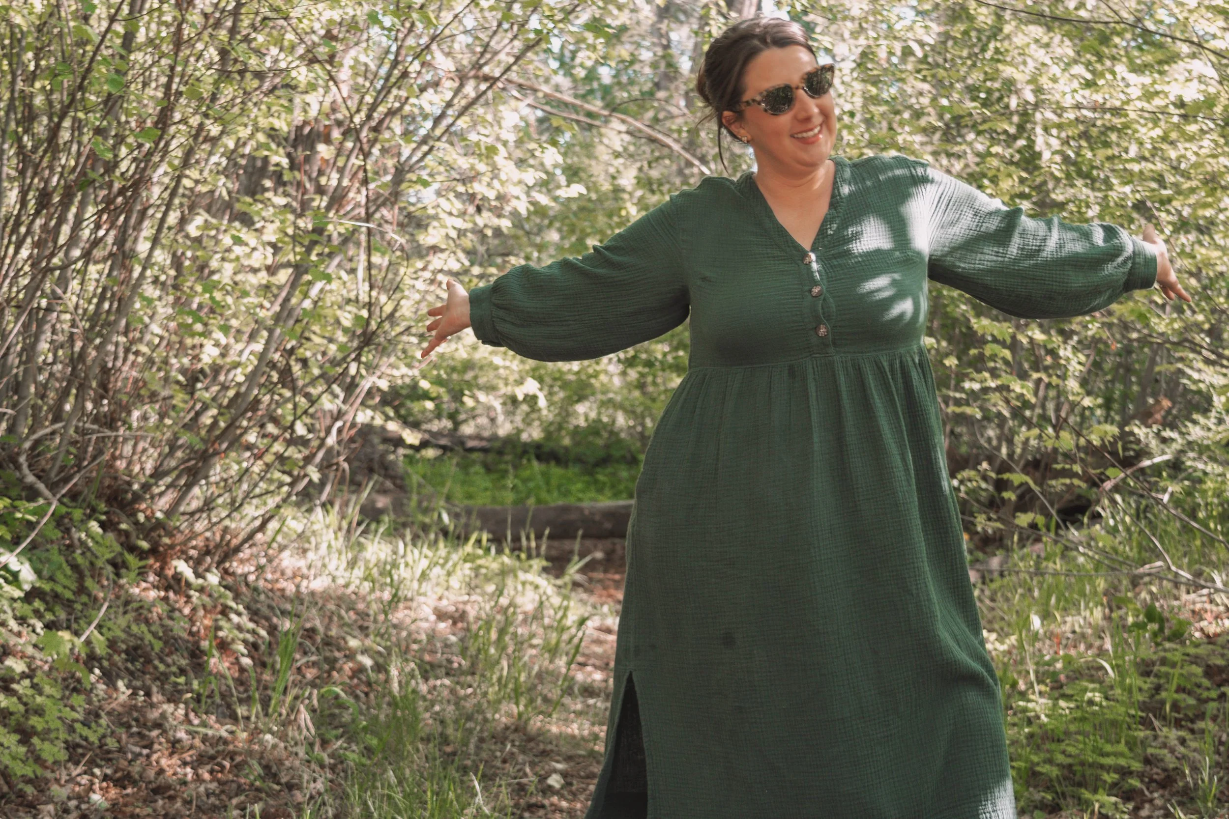 A woman with dark hair wearing sunglasses and a green dress with puffed sleeves standing in a wooded area with her arms outstretched and smiling.