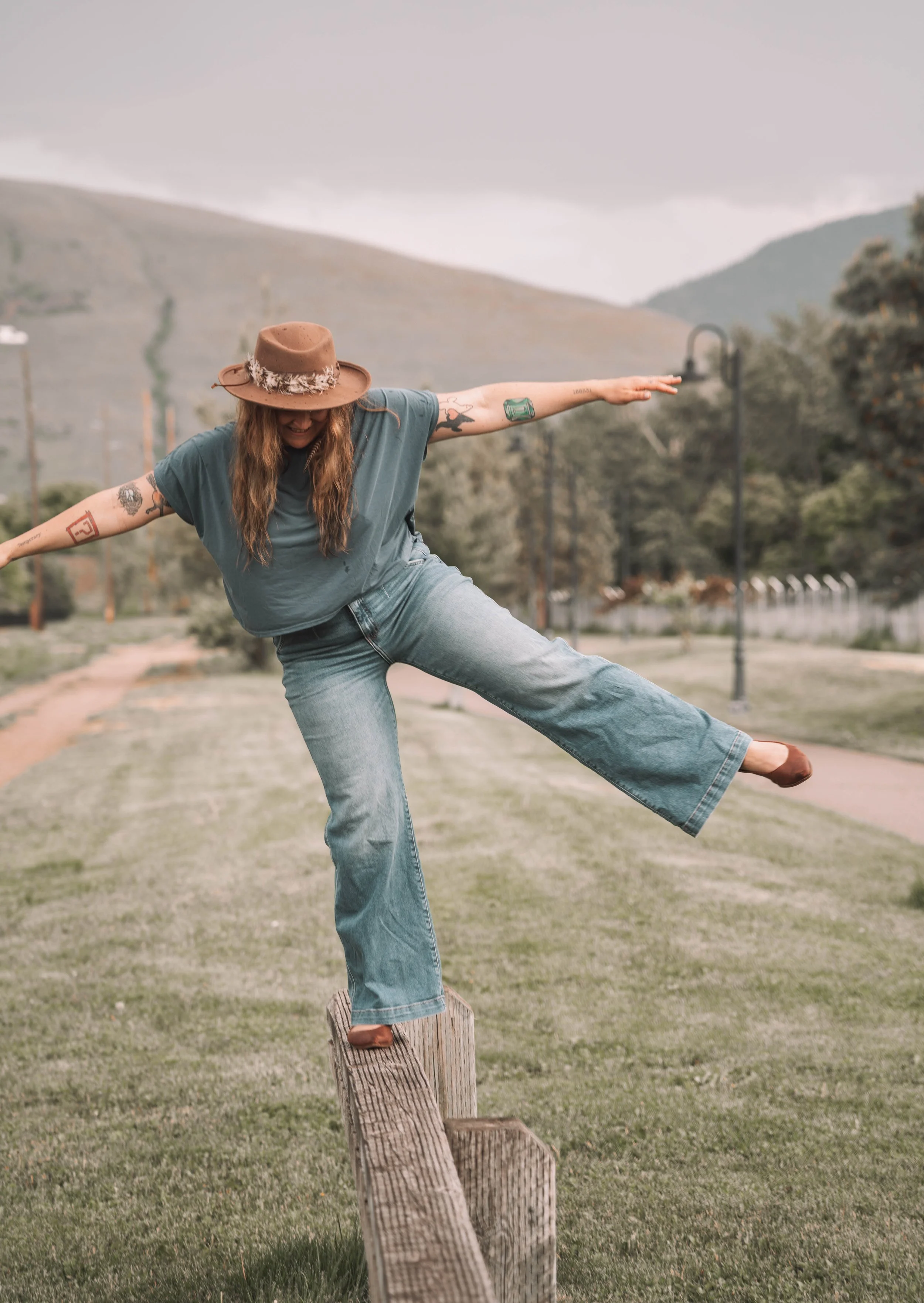 Woman with long hair and tattoos balancing on a wooden post, with hills and trees in the background on a cloudy day.
