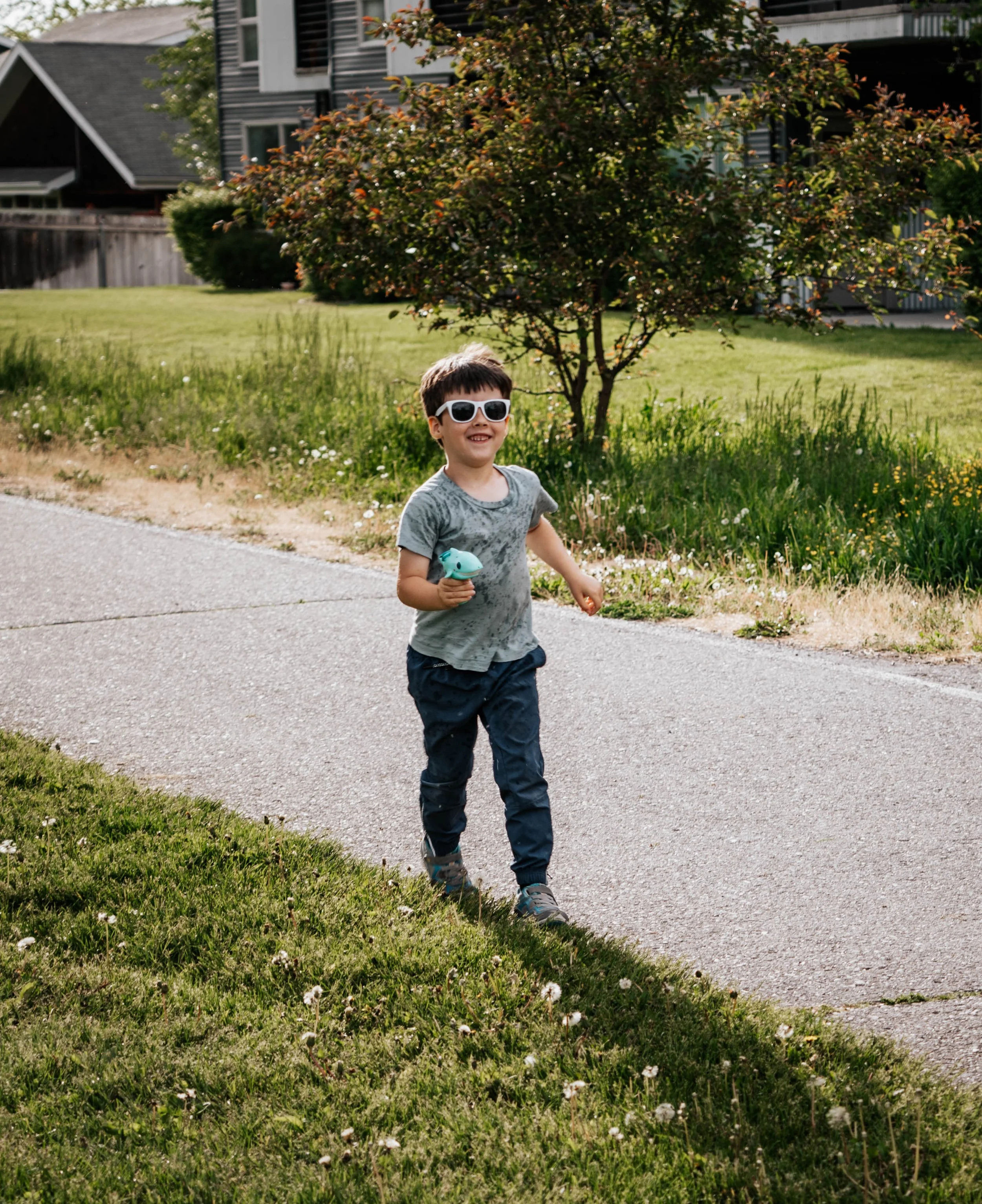 A young boy with short brown hair, wearing dark sunglasses, a gray shirt, and dark pants, happily running in a grassy yard while holding a teal toy in his right hand, with a tree and a house in the background.