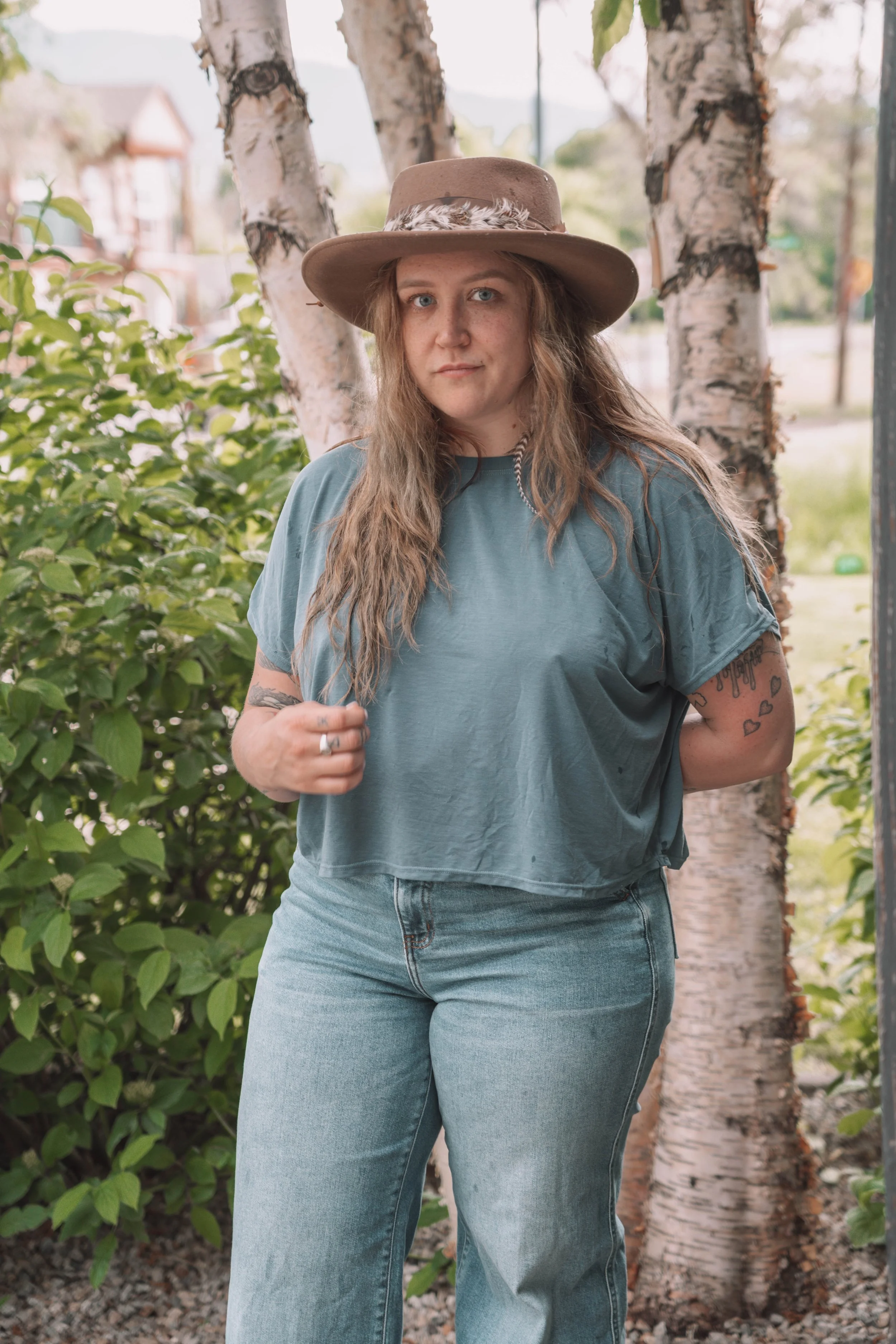 A woman with long wavy hair, wearing a beige hat with a feather, light blue T-shirt, and blue jeans, standing outdoors near trees and bushes.