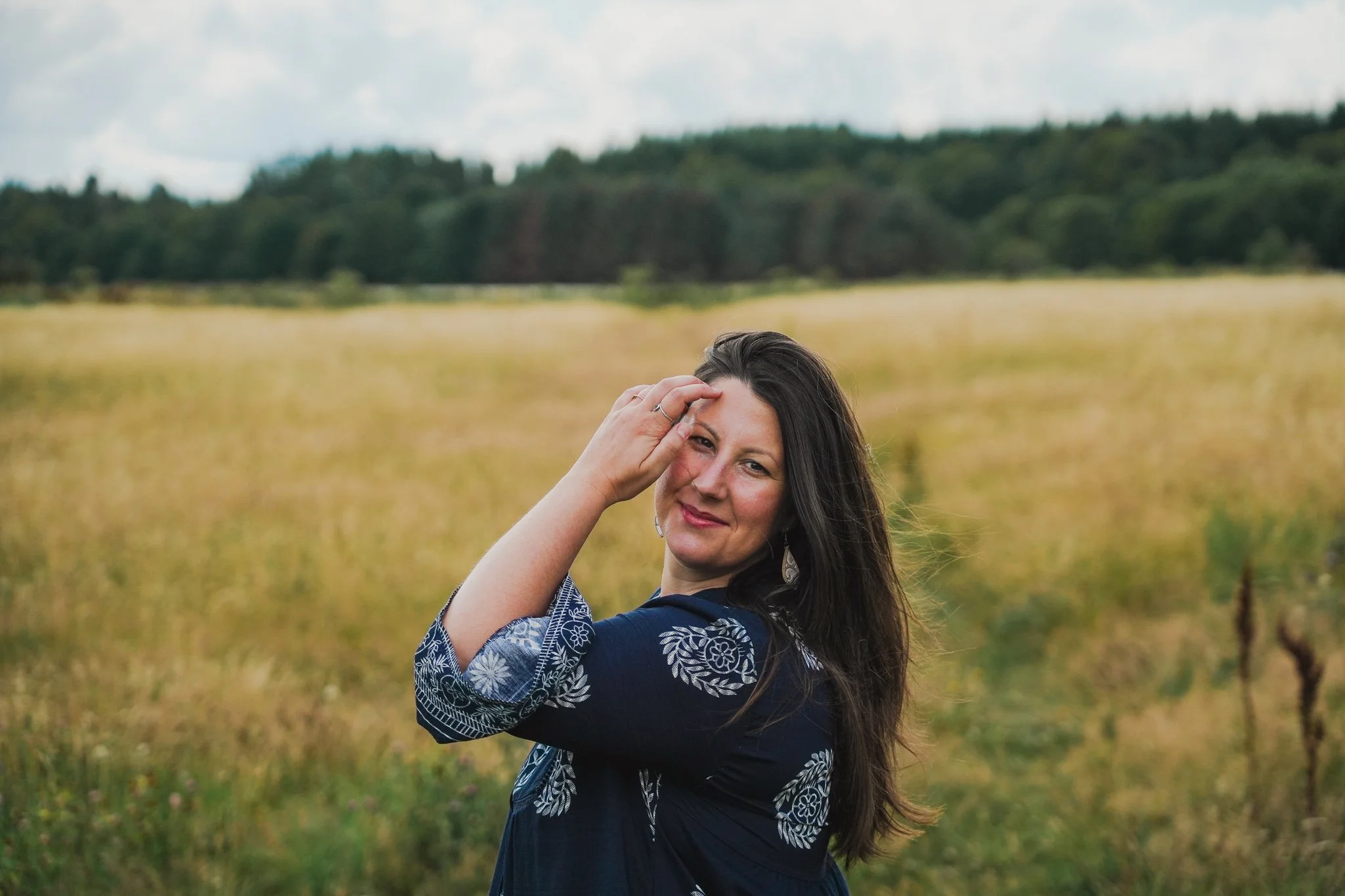 A woman with long dark hair outdoors in a field with a forest in the background, tilting her head and smiling, wearing a blue patterned top.