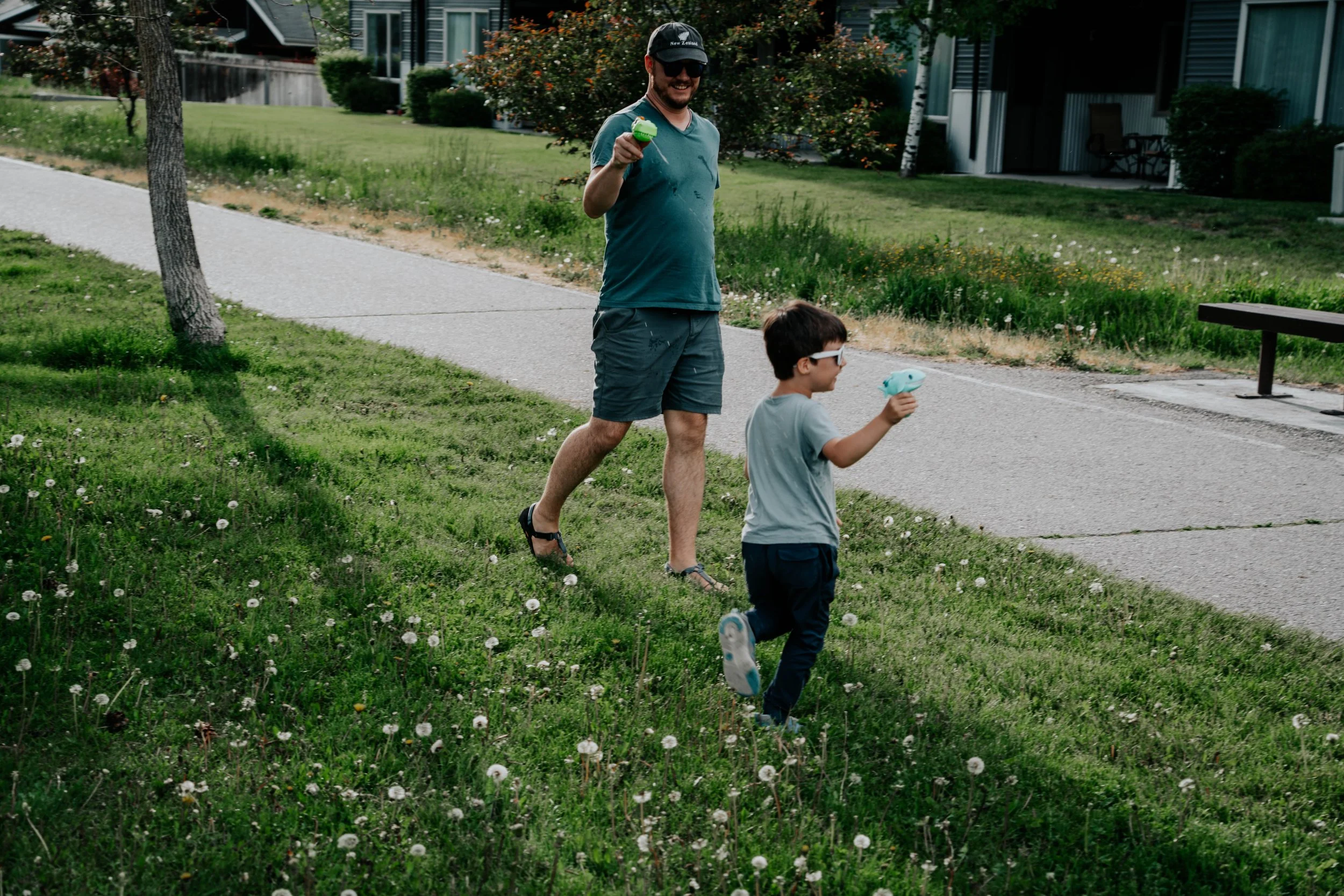 A man and a young boy walking outdoors in a green neighborhood park, holding toy water guns, with houses and trees in the background.