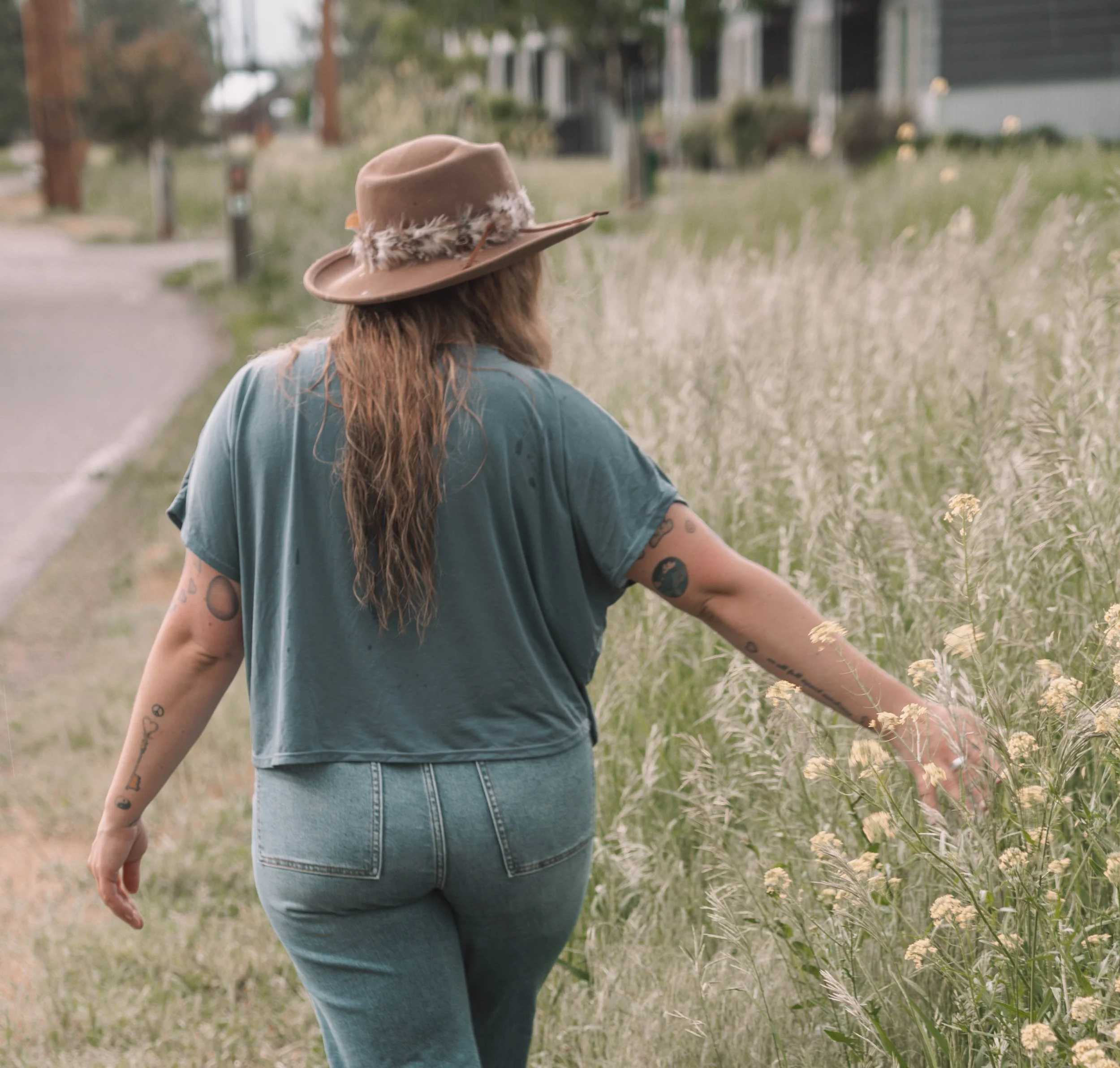 A woman with long hair and tattoos on her arms is walking through a grassy field, wearing a blue shirt and a brown hat with a feather detail, during daytime.