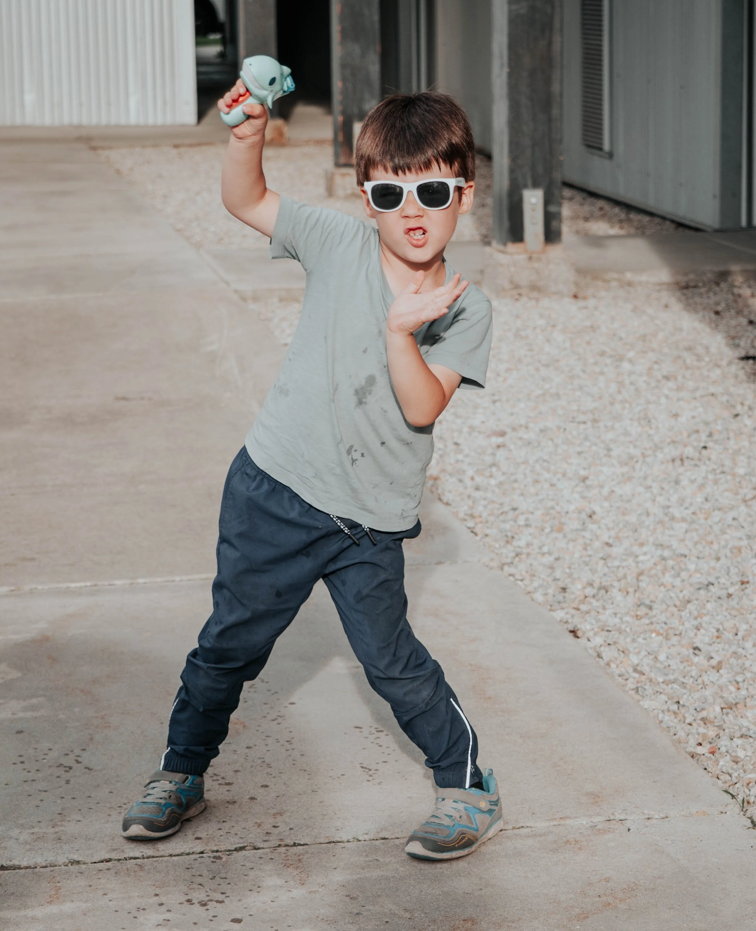 Young boy wearing white sunglasses, gray t-shirt, and dark pants, striking a playful pose with one hand on his chest and the other raised holding a toy, outdoors on a concrete sidewalk.