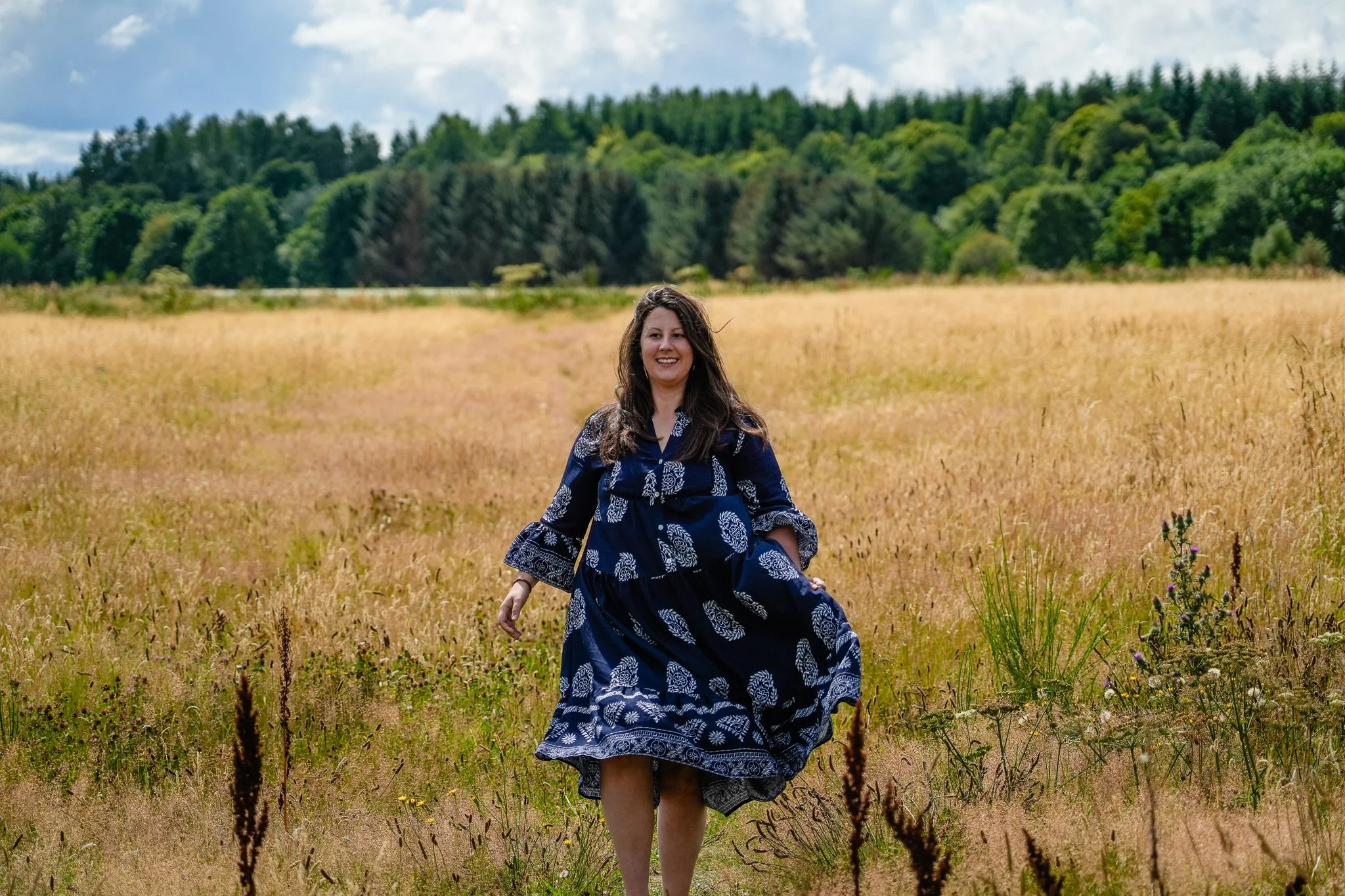 A woman with long brown hair, wearing a blue dress with white leaf patterns, is smiling and walking through a yellow-green field with tall grass and wildflowers, surrounded by green trees and hills under a partly cloudy sky.