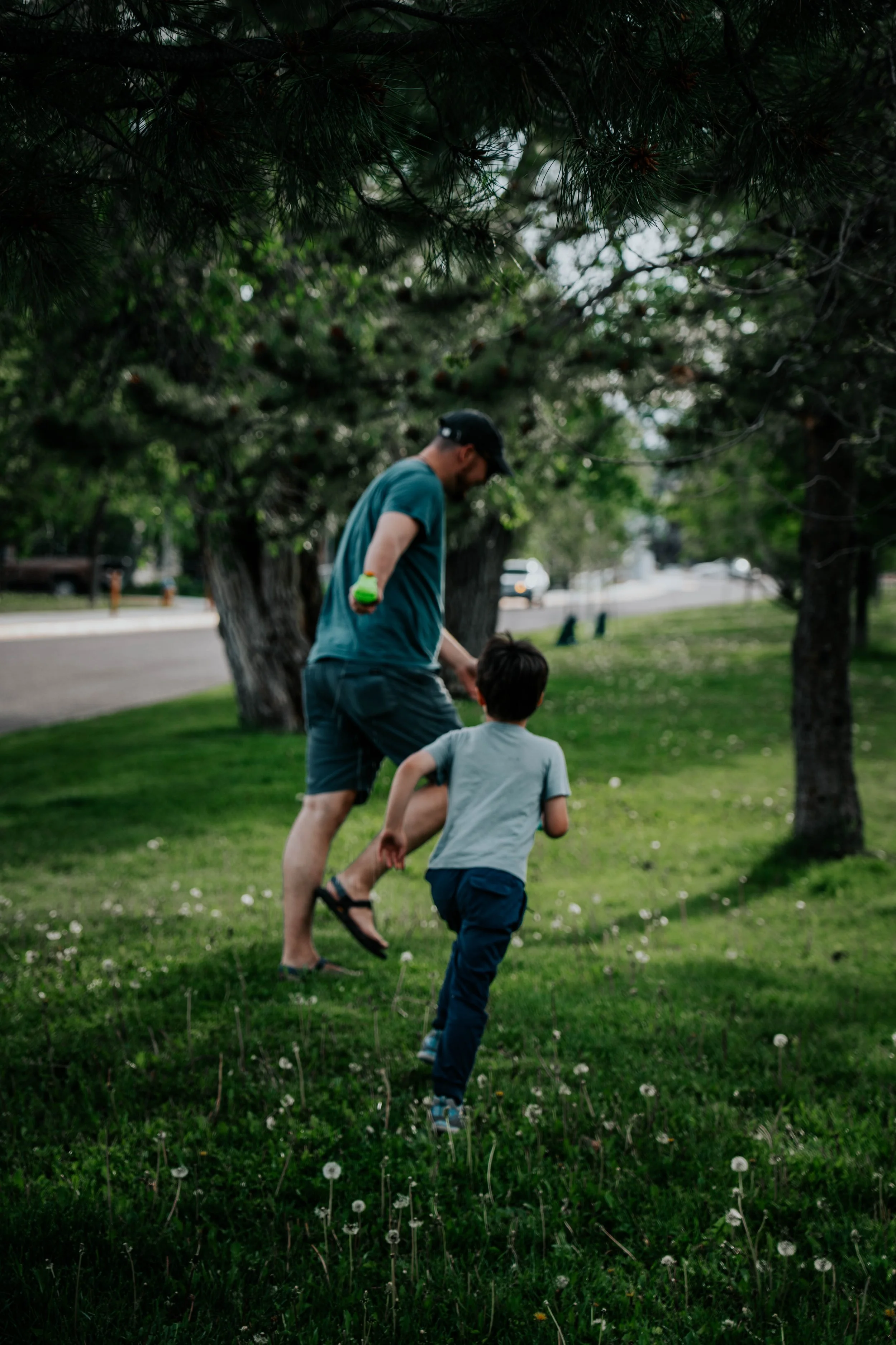 An adult male and a young boy playing outdoors in a grassy area with trees. The adult is holding the boy's hand as they run or walk together, with the adult wearing a cap, t-shirt, and shorts, and the boy wearing a t-shirt and pants.