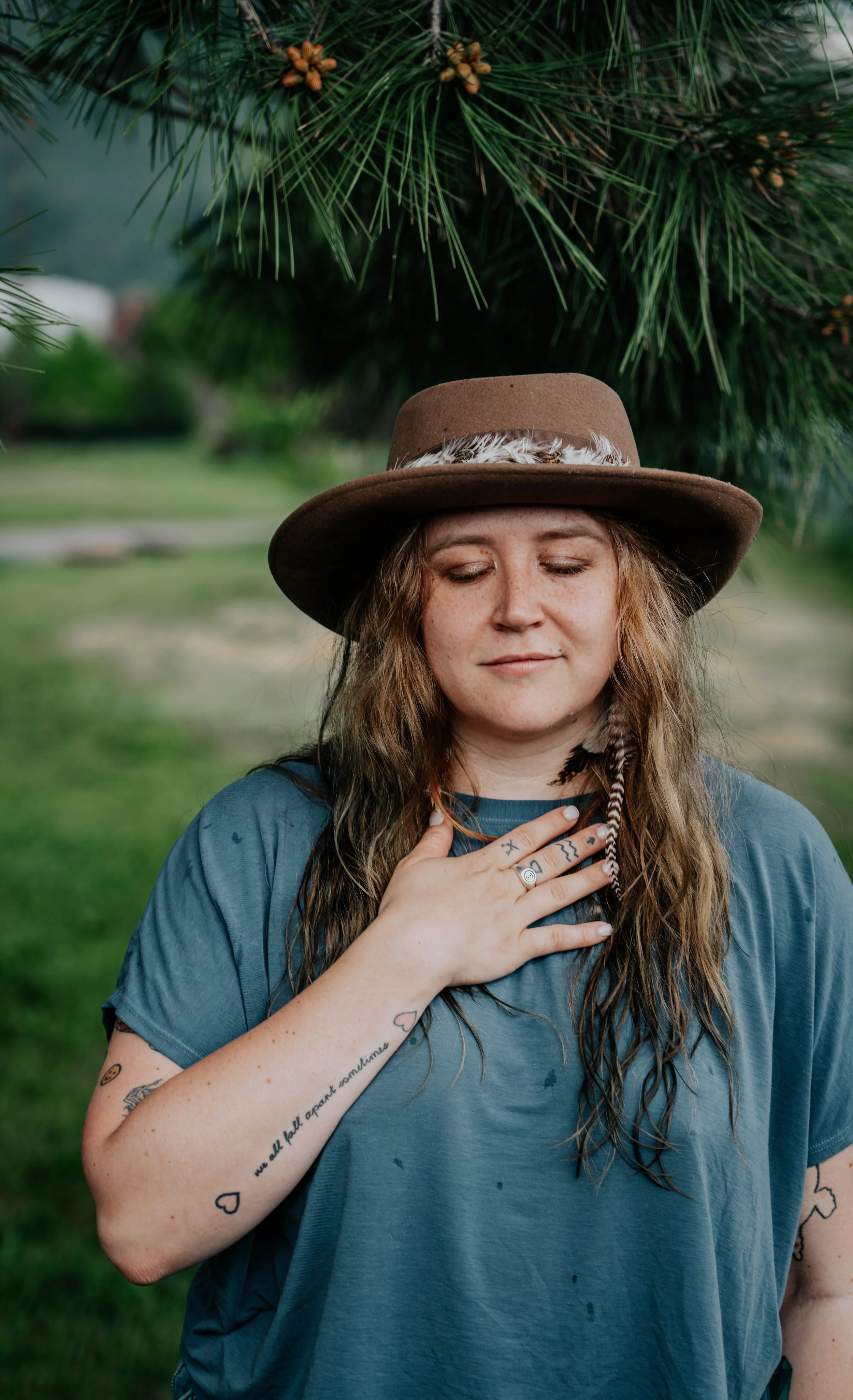 A woman with long hair, tattoos, and rings on her fingers, wearing a wide-brimmed hat with feathers, and a blue T-shirt, standing outdoors near pine trees, with her eyes closed and hand on her chest.