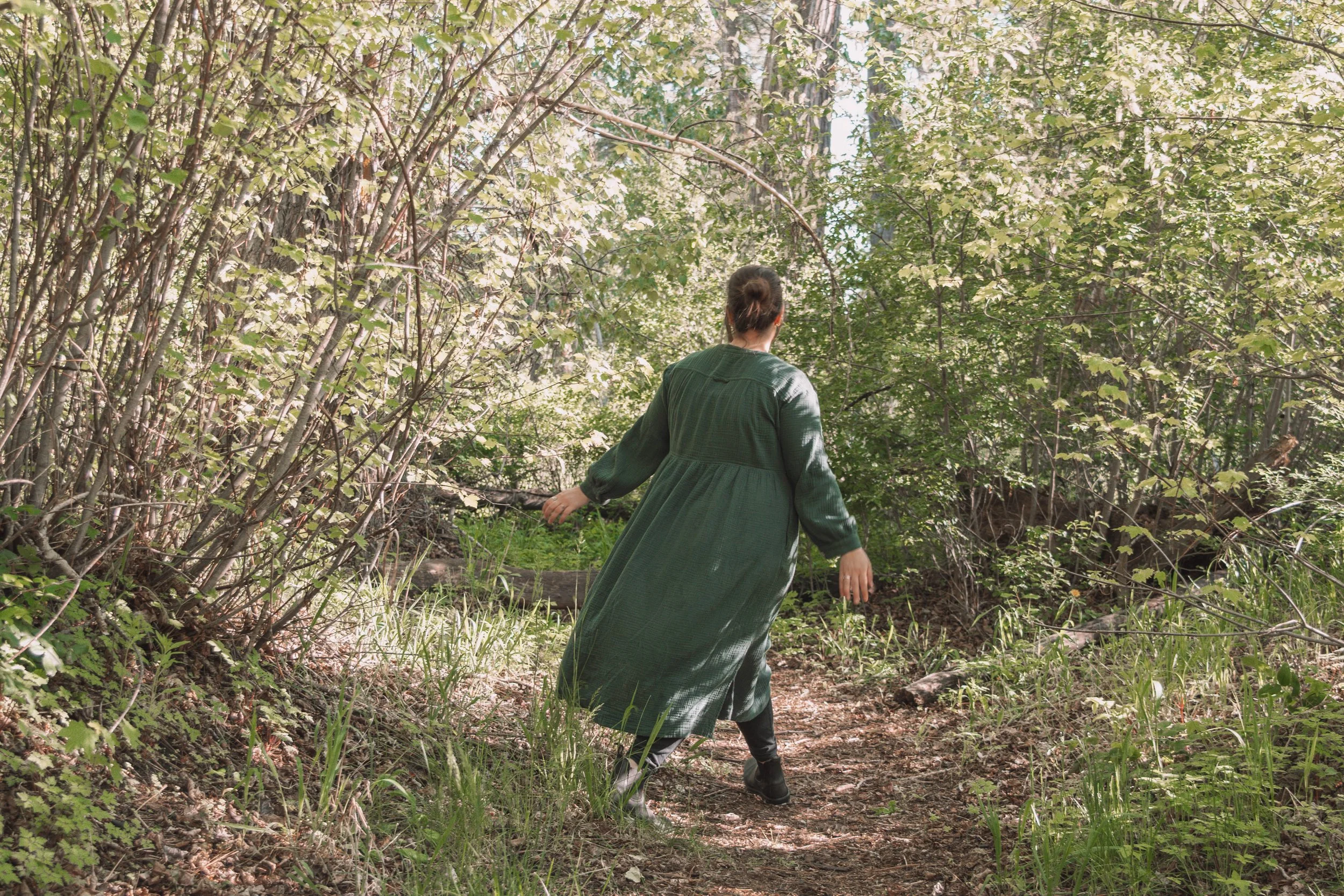A woman in a green dress walking through a dense, sunlit forest with lush green leaves and tall trees.