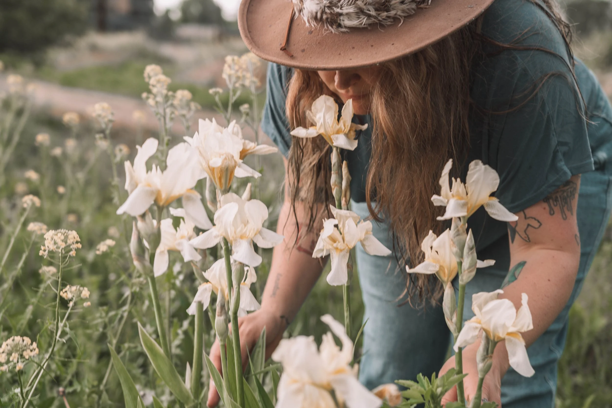 A woman with long hair wearing a hat and a blue shirt is in a garden, smelling or examining white iris flowers.