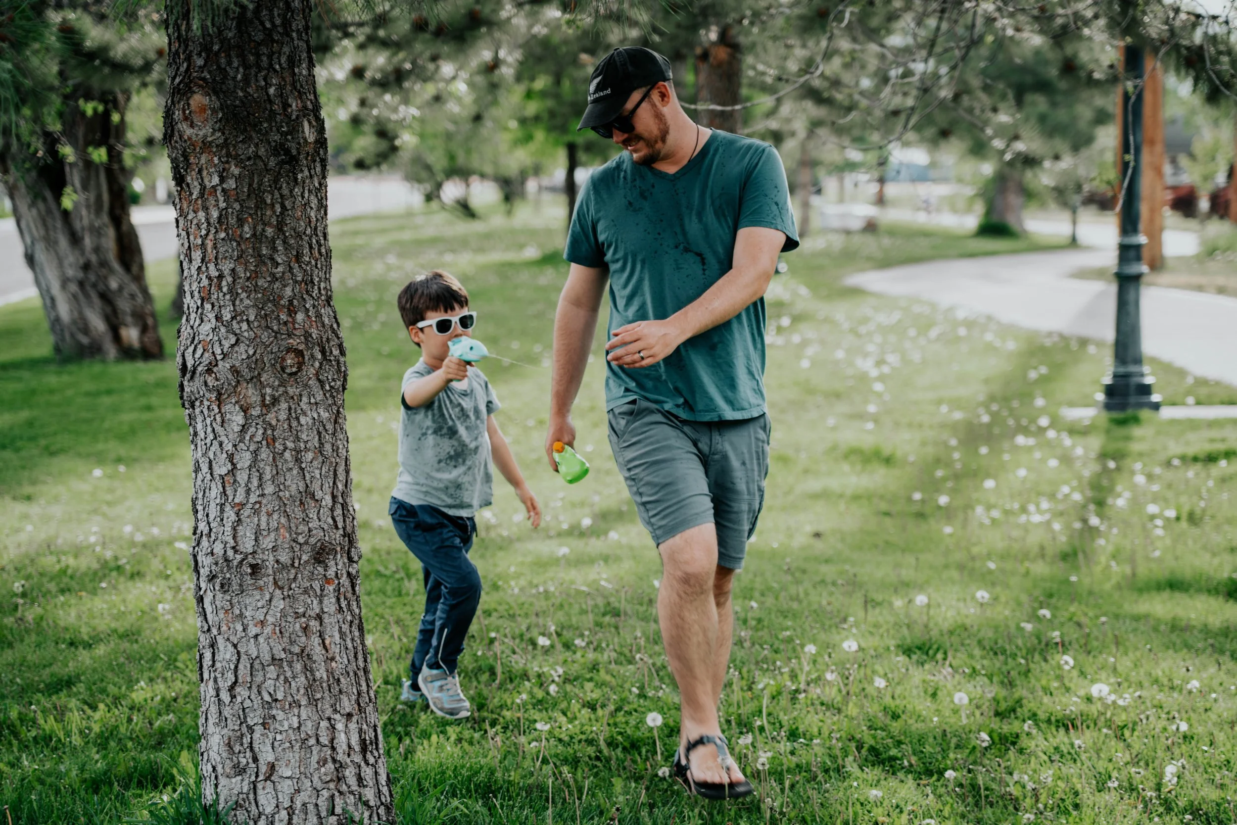 A man and a young boy walking in a park on a sunny day. The boy is holding a blue toy and wearing sunglasses, while the man is wearing a cap, sunglasses, and casual clothing. They are walking on a grassy area with trees and a sidewalk in the backgrou
