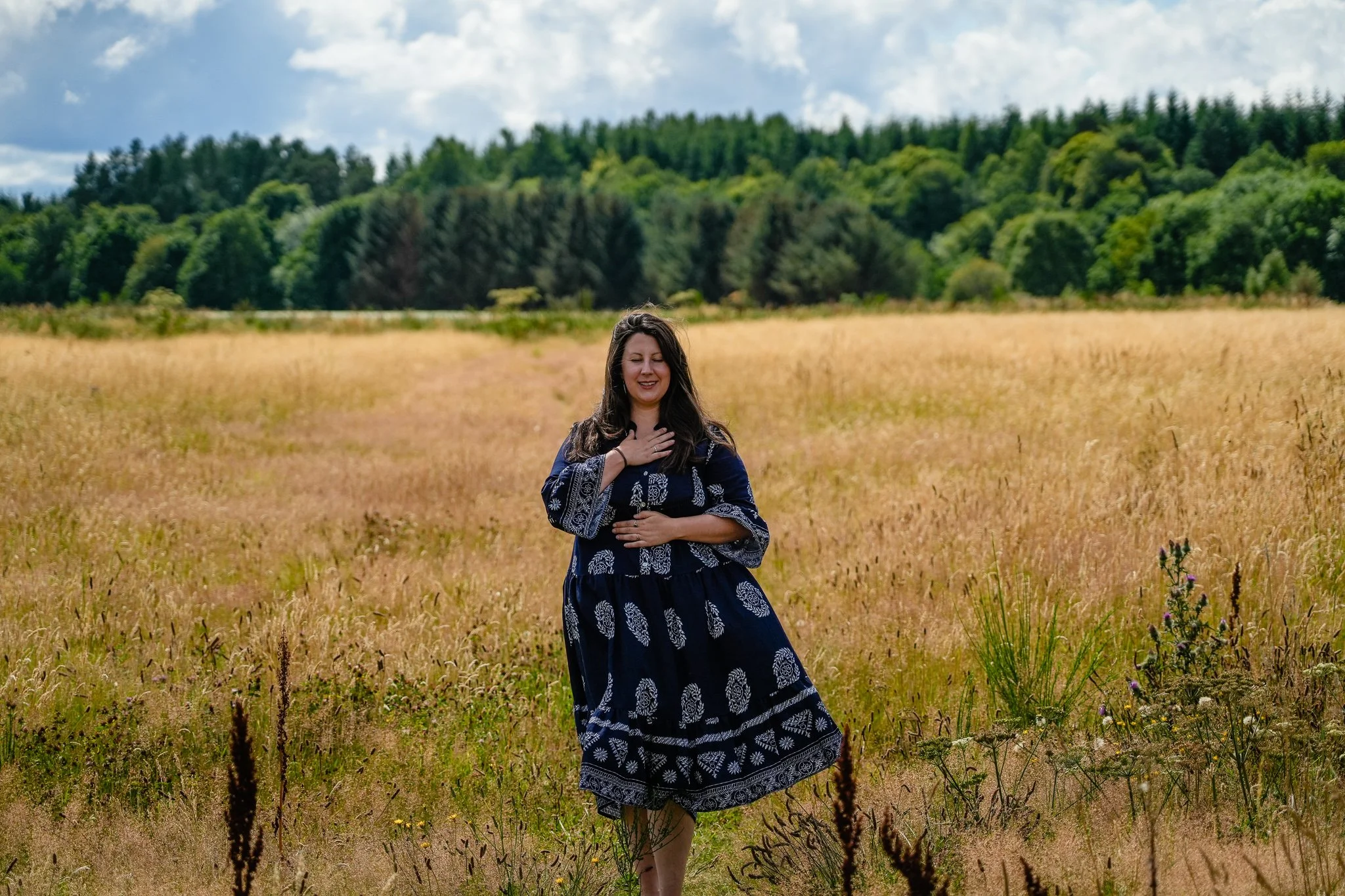 A woman standing in a field of tall grass with a forested hill and partly cloudy sky in the background, smiling with eyes closed and hand over her chest.