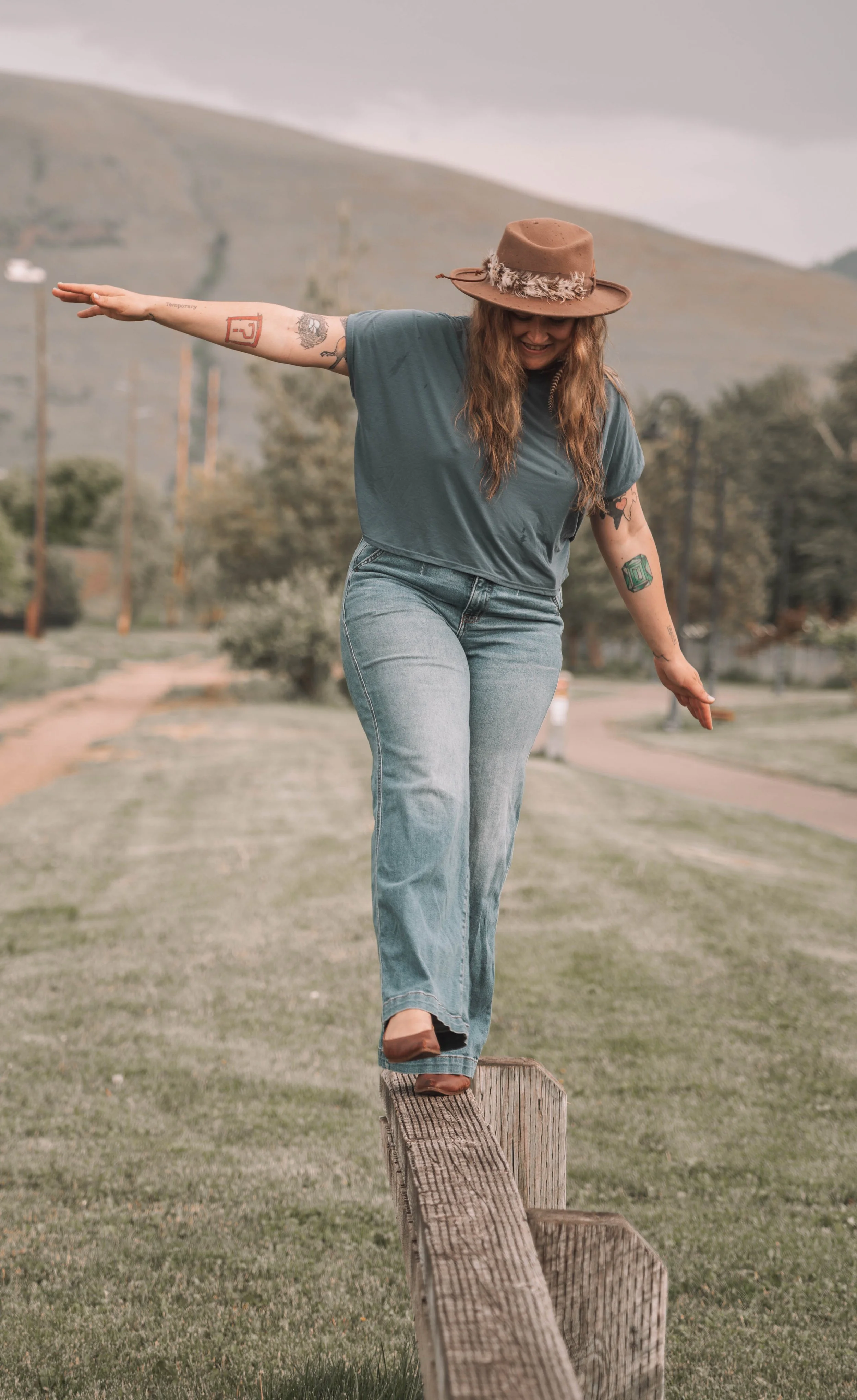 A woman with long hair, tattoos, and a hat balancing on a wooden rail outdoors with hills and trees in the background.