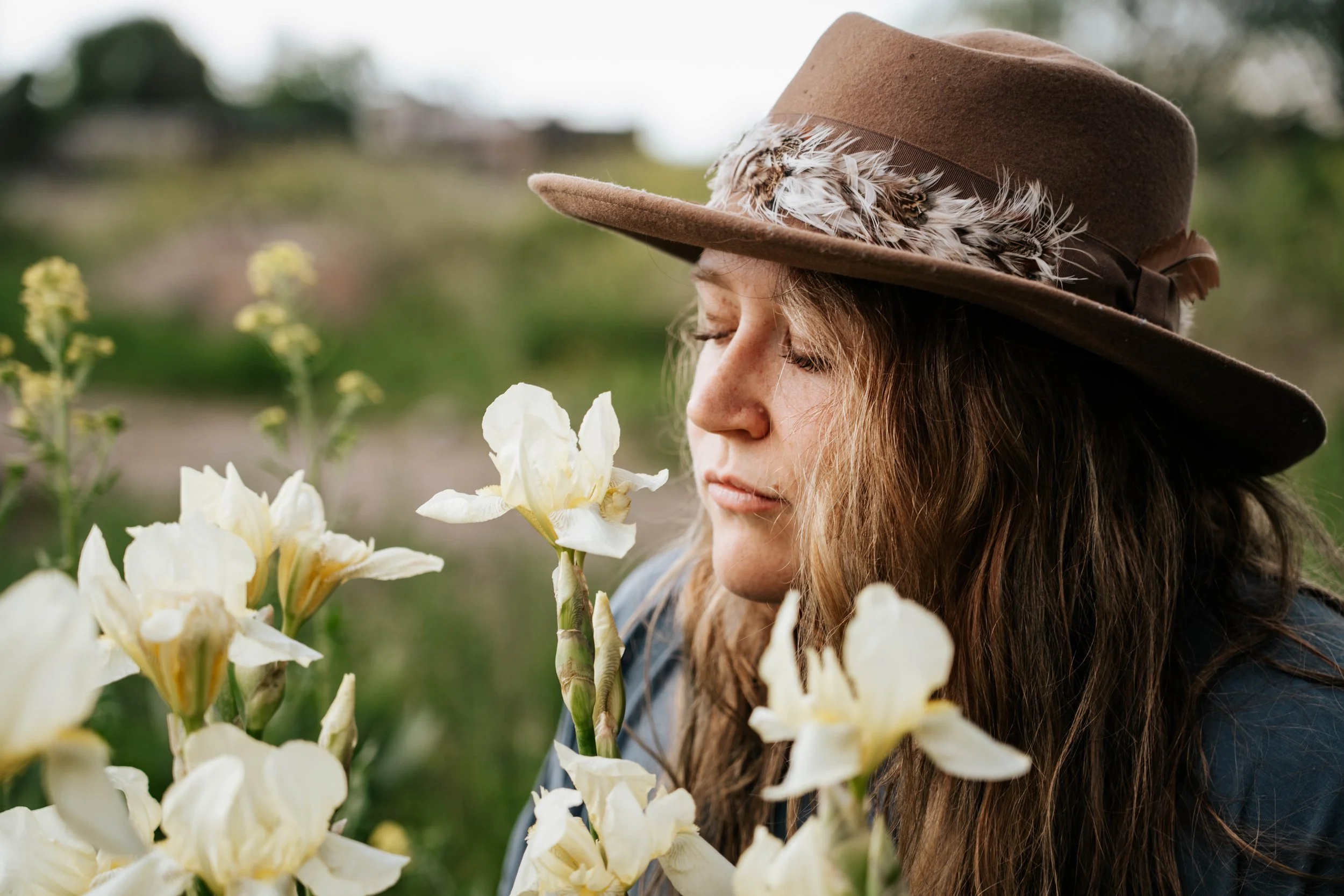 A woman with long hair wearing a brown hat with feathers is smelling white flowers in a garden.