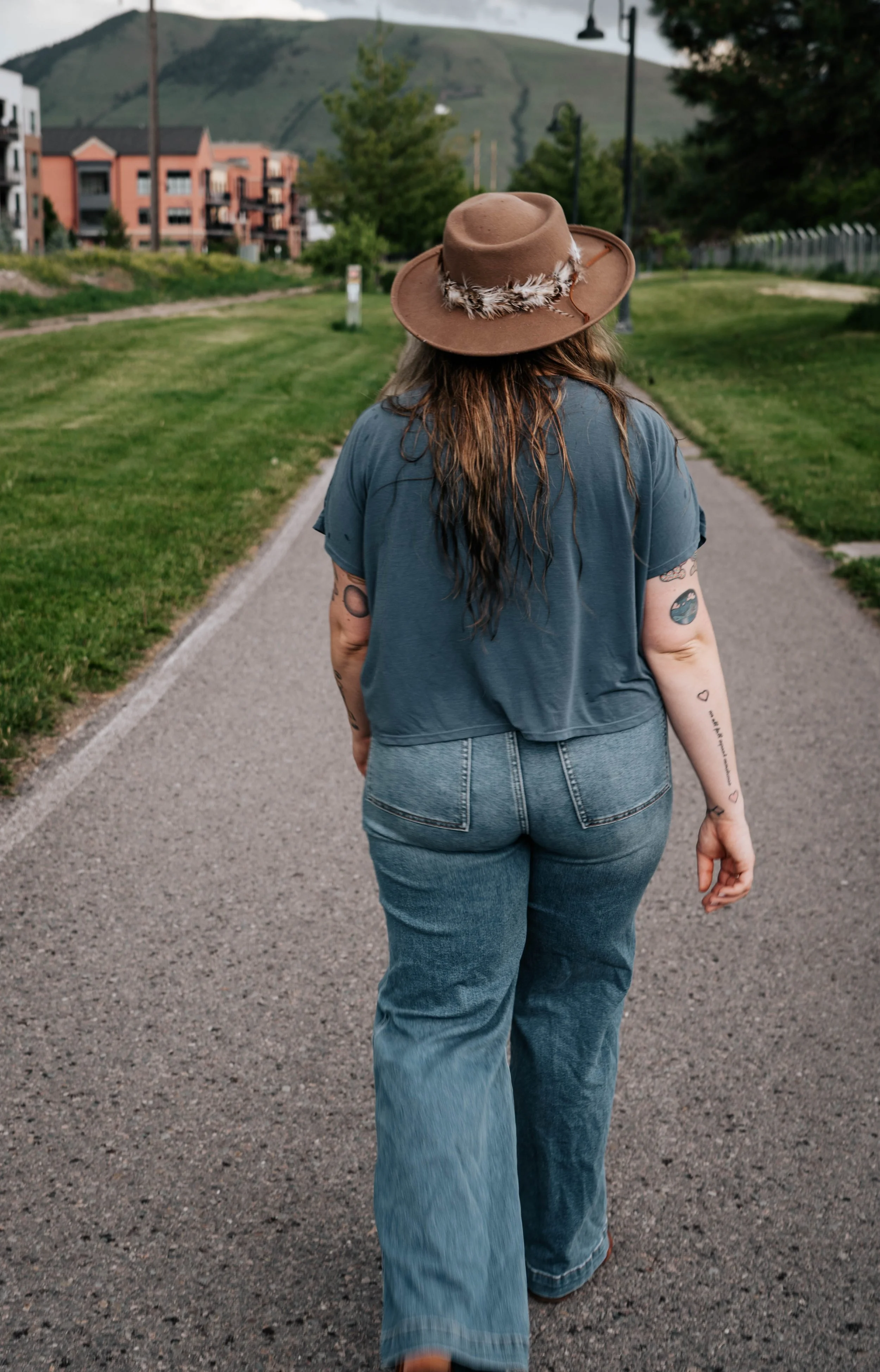Person walking on a paved path with long wavy hair, wearing a tan wide-brimmed hat with feather details, a blue t-shirt, and high-waisted jeans, with a grassy area and buildings in the background.