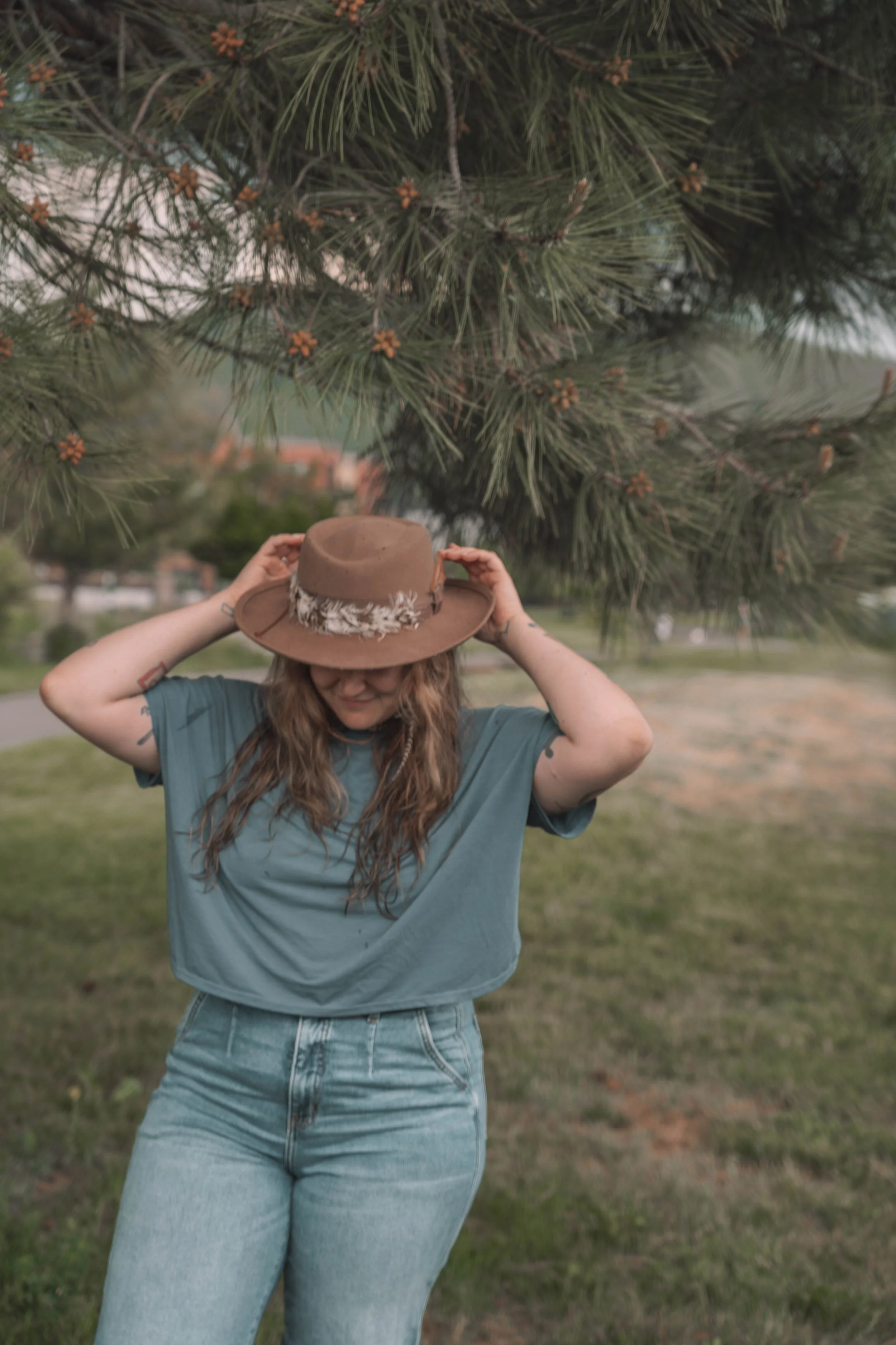 A woman with long wavy hair wearing a teal t-shirt, light blue jeans, and a brown wide-brimmed hat with feathers, standing outdoors under a pine tree.
