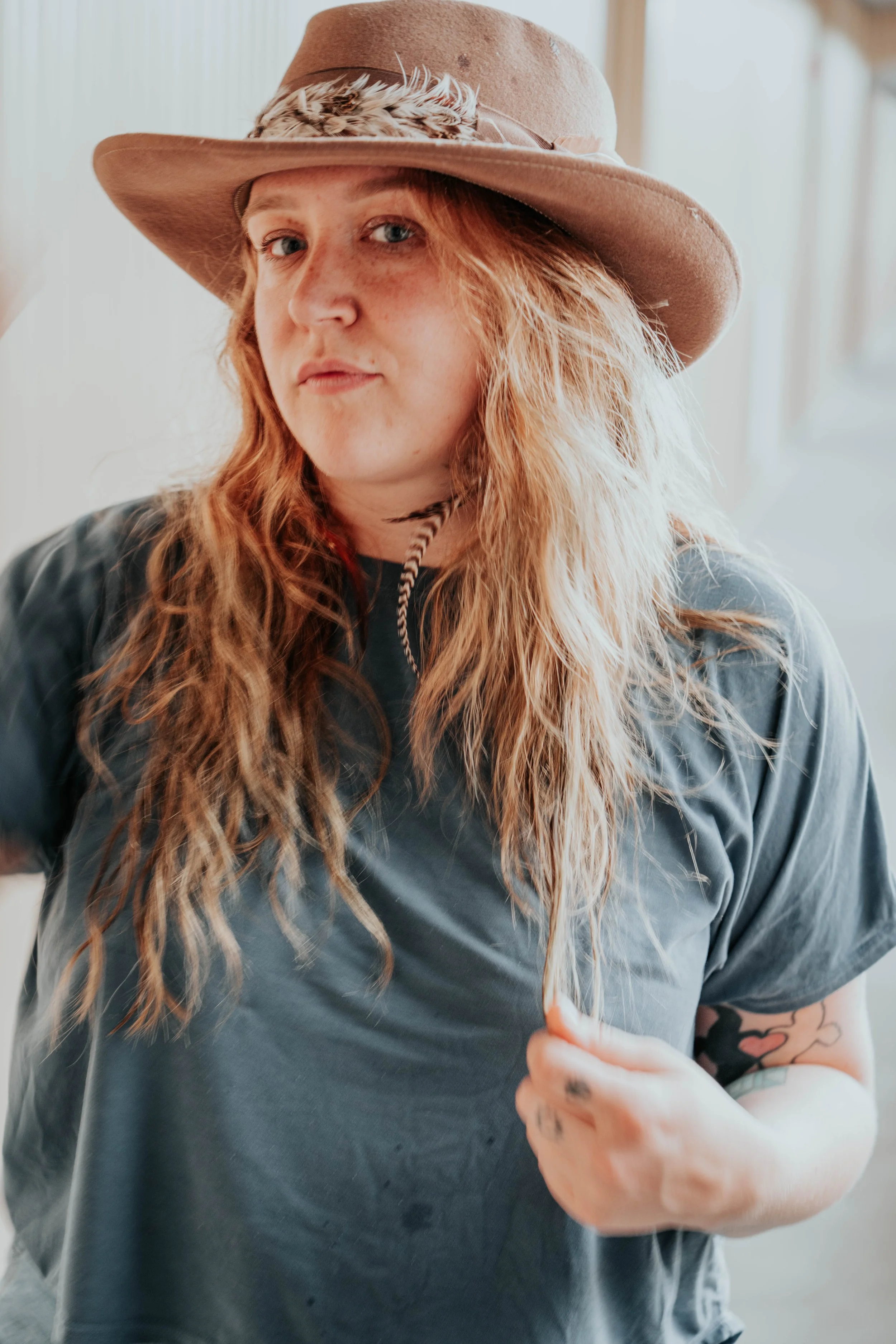 A woman with long, wavy red hair wearing a wide-brimmed, tan hat with feathers, a gray T-shirt, and visible tattoos on her arm, standing indoors near a large window.