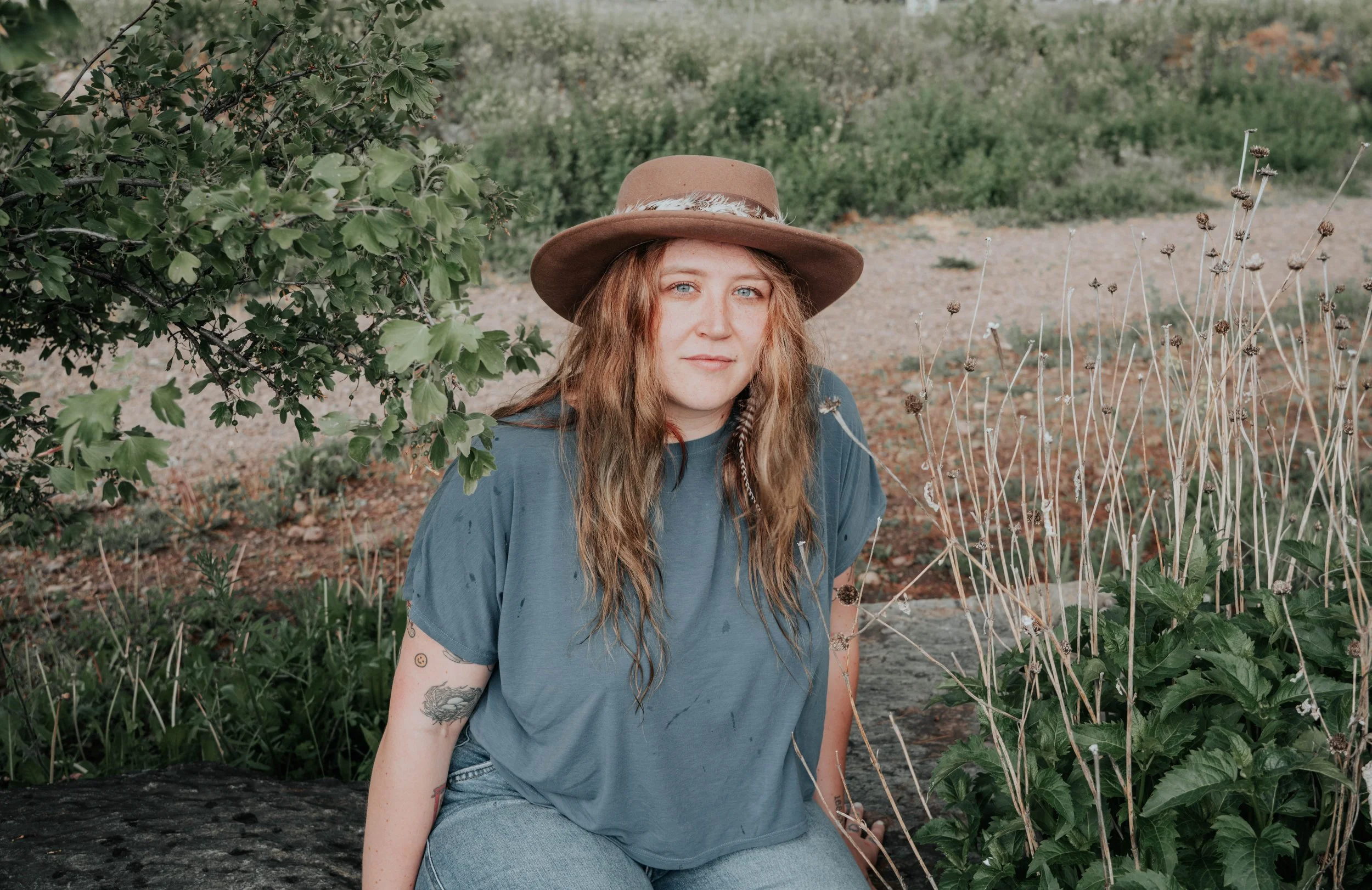 A woman with long wavy hair wearing a brown wide-brimmed hat and a blue t-shirt, sitting outdoors near green plants and dried flowers.