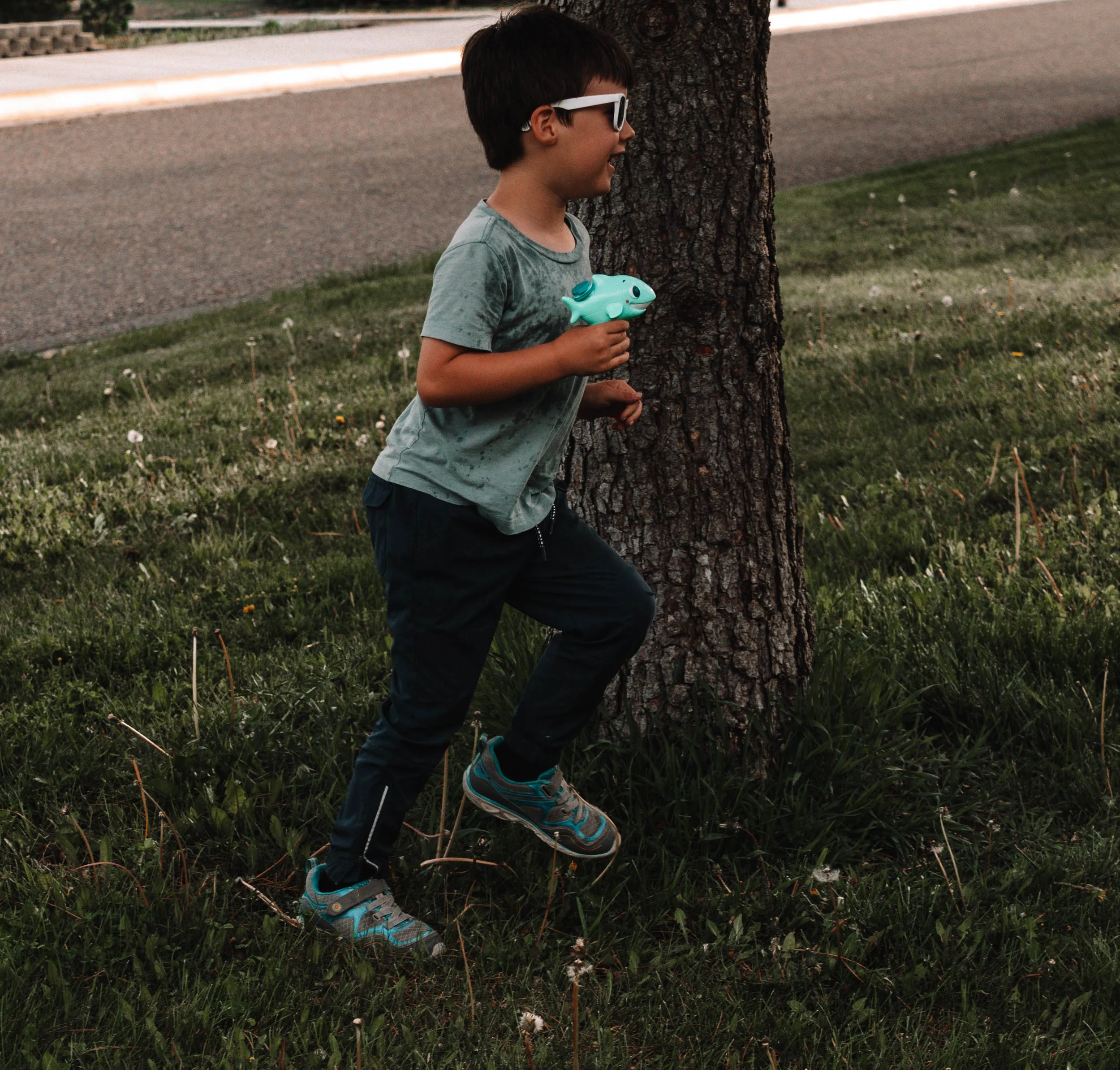 A young boy running outdoors on a grassy area near a tree, holding a blue toy fish, wearing sunglasses, a damp gray T-shirt, dark pants, and sneakers.