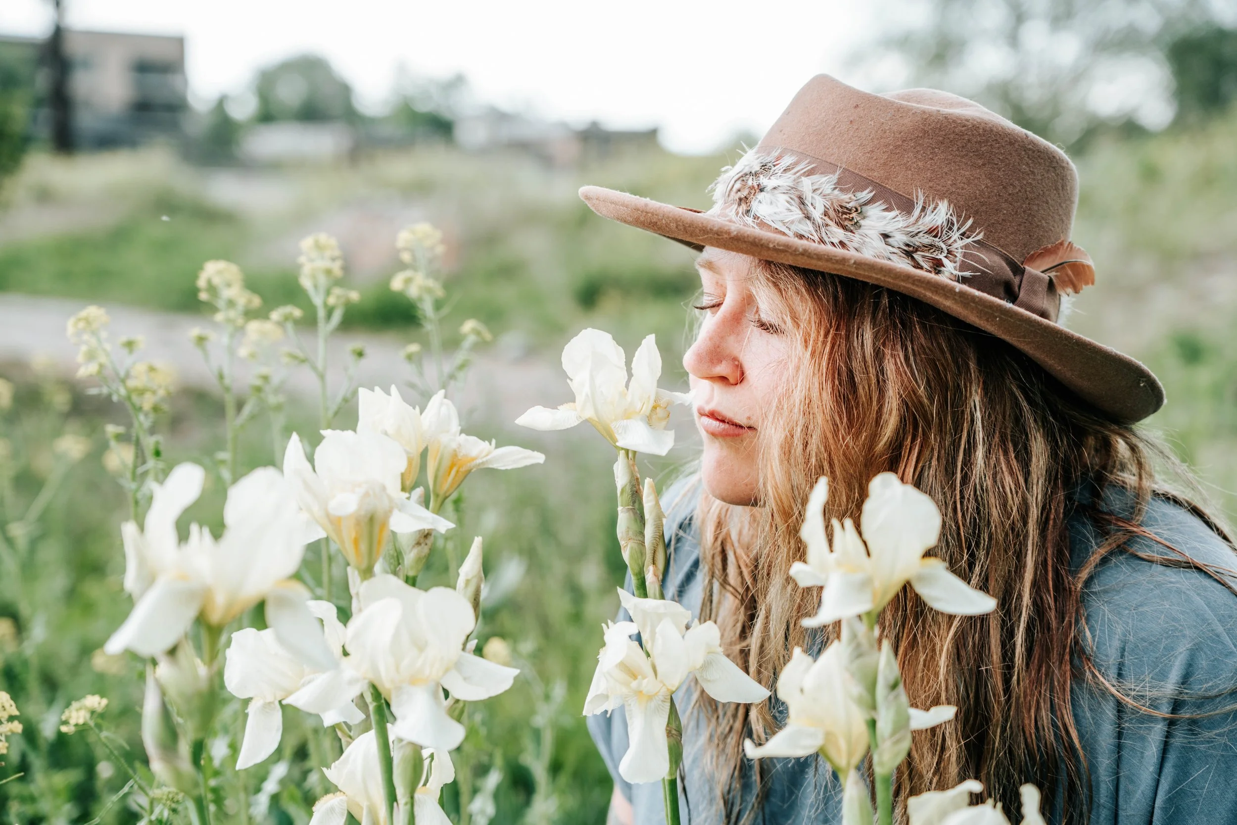 A woman with long hair and a tan felt hat decorated with feathers smelling white flowers in a garden.