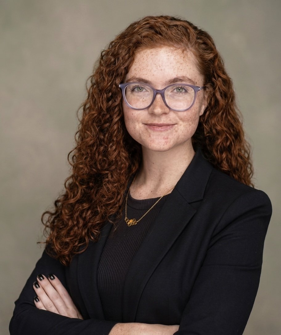 A woman with long, curly red hair and glasses, wearing a black blazer and a gold necklace, posing with arms crossed against a plain background.
