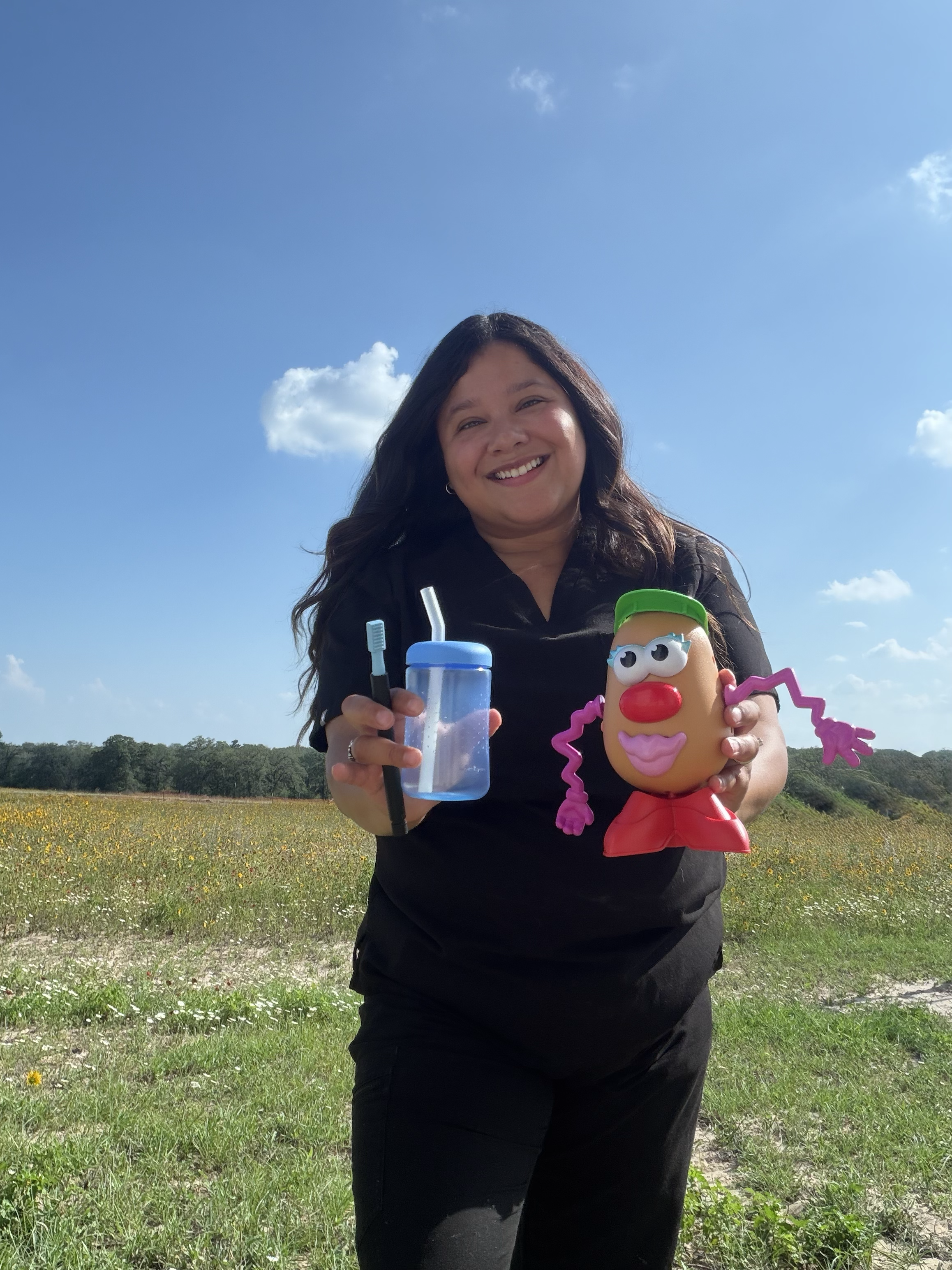 Smiling woman outdoors holding a toy character, sunlit field and blue sky in the background.