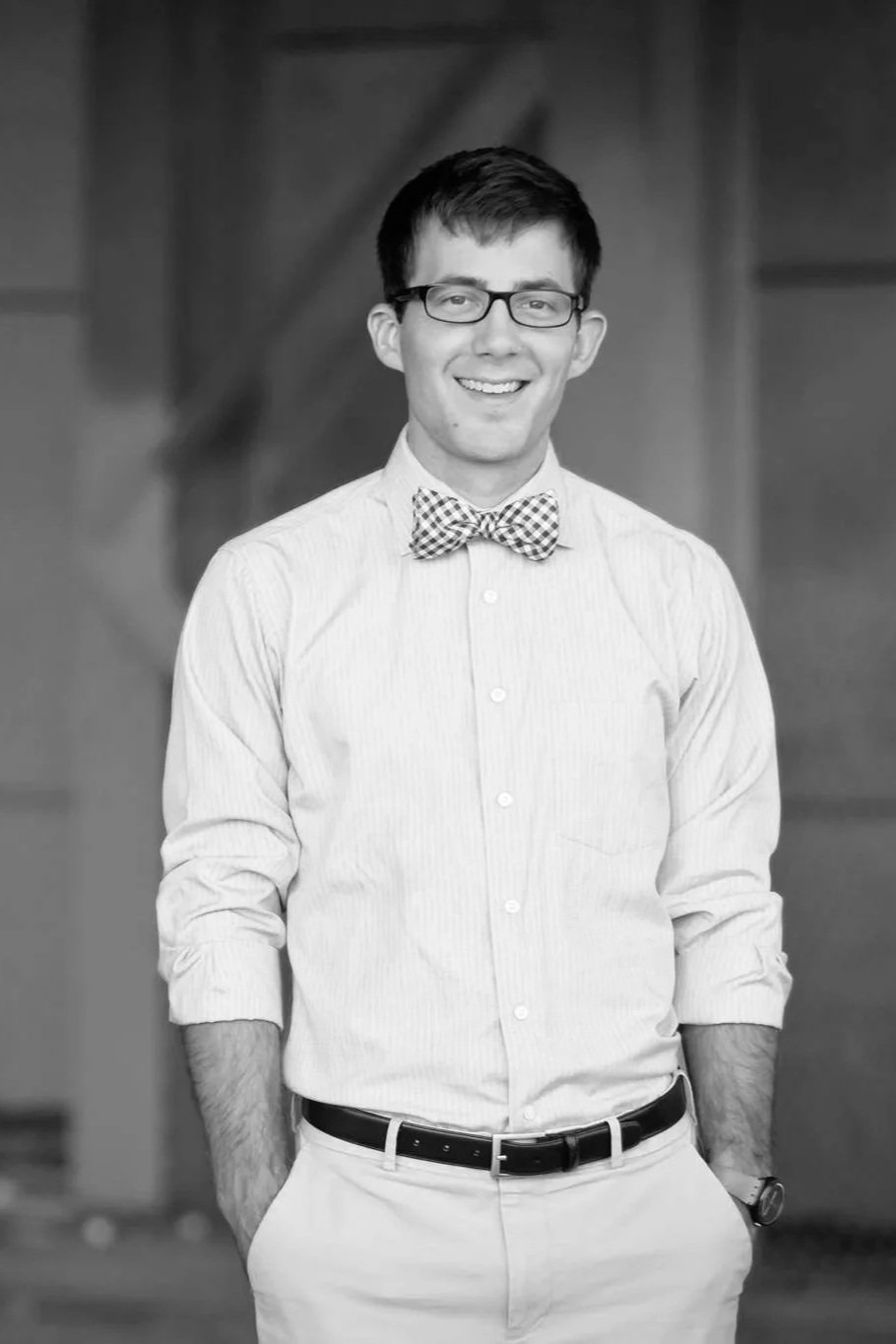 A young man with glasses, smiling, wearing a light-colored button-up shirt, a patterned bow tie, light-colored pants, and a belt, standing indoors.