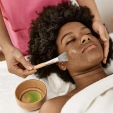 Woman receiving a facial treatment, with a skincare product being applied to her face by a professional, and a bowl with green substance nearby.