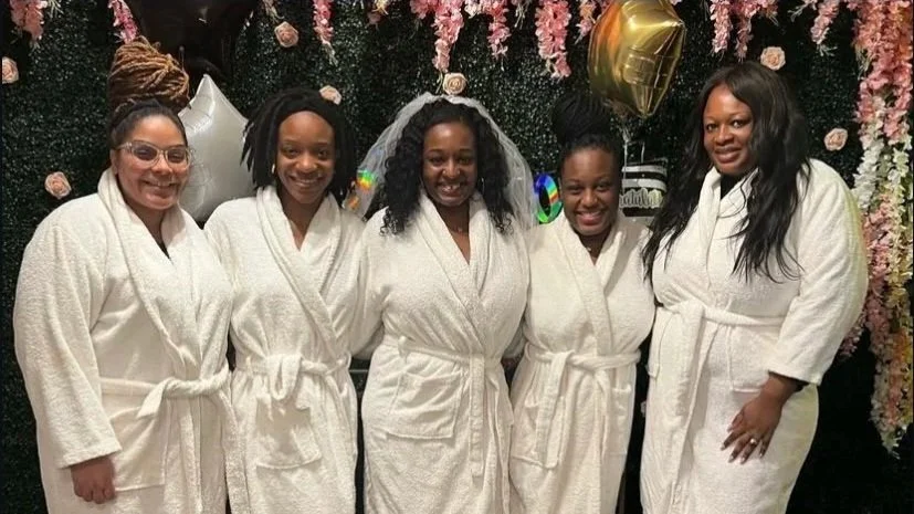 Five women in white bathrobes standing in front of a floral backdrop with pink flowers and balloons, smiling at the camera.