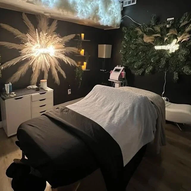 Salon room with a massage table covered in gray and black sheets, decorative plants on the wall, candles on shelves, and a fireworks display projected on the wall.