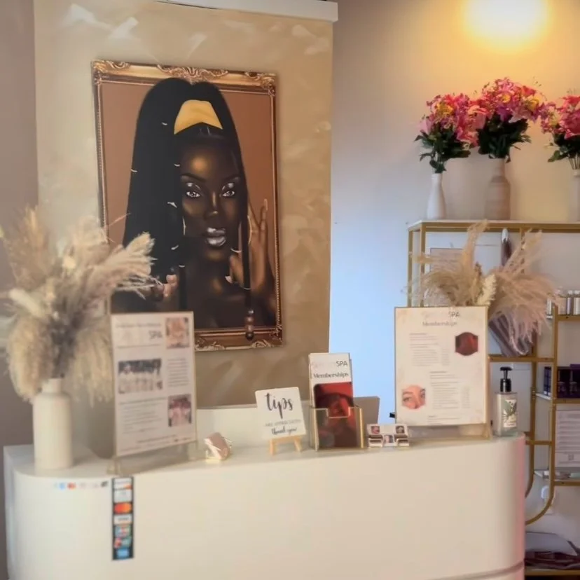 Interior of a spa reception with a large framed portrait of a woman with dark skin and hair styled with a yellow feather accessory. The reception desk has informational signs, tips, and promotional material. Decor includes dried pampas grass and fresh pink flowers in vases.