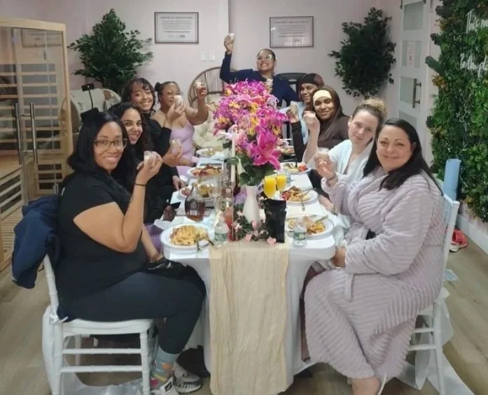 A group of nine women sitting around a dining table, celebrating with food and drinks. There is a large floral centerpiece, and they are smiling and raising their glasses in a toast.