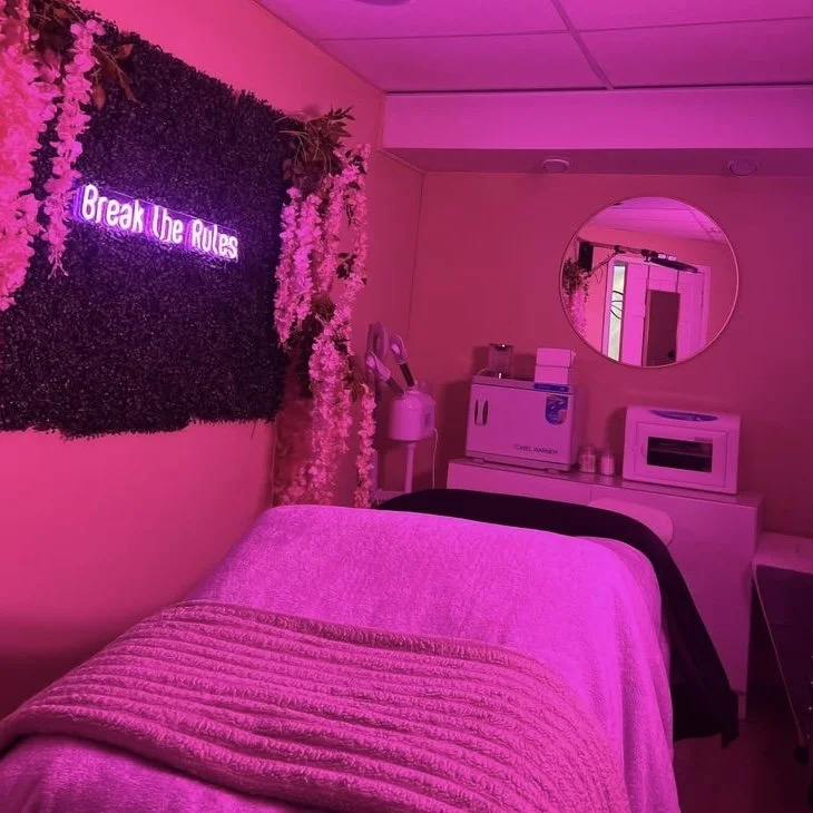A room with pink lighting and a bed covered with a quilt. On the wall, a sign says 'Break the Rules' and is lit with purple neon. There are decorative flowers and plants, a mirror, and some microwave and beverage station appliances in the background.