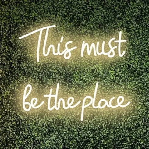 Neon sign with the phrase 'This must be the place' against a background of green foliage.