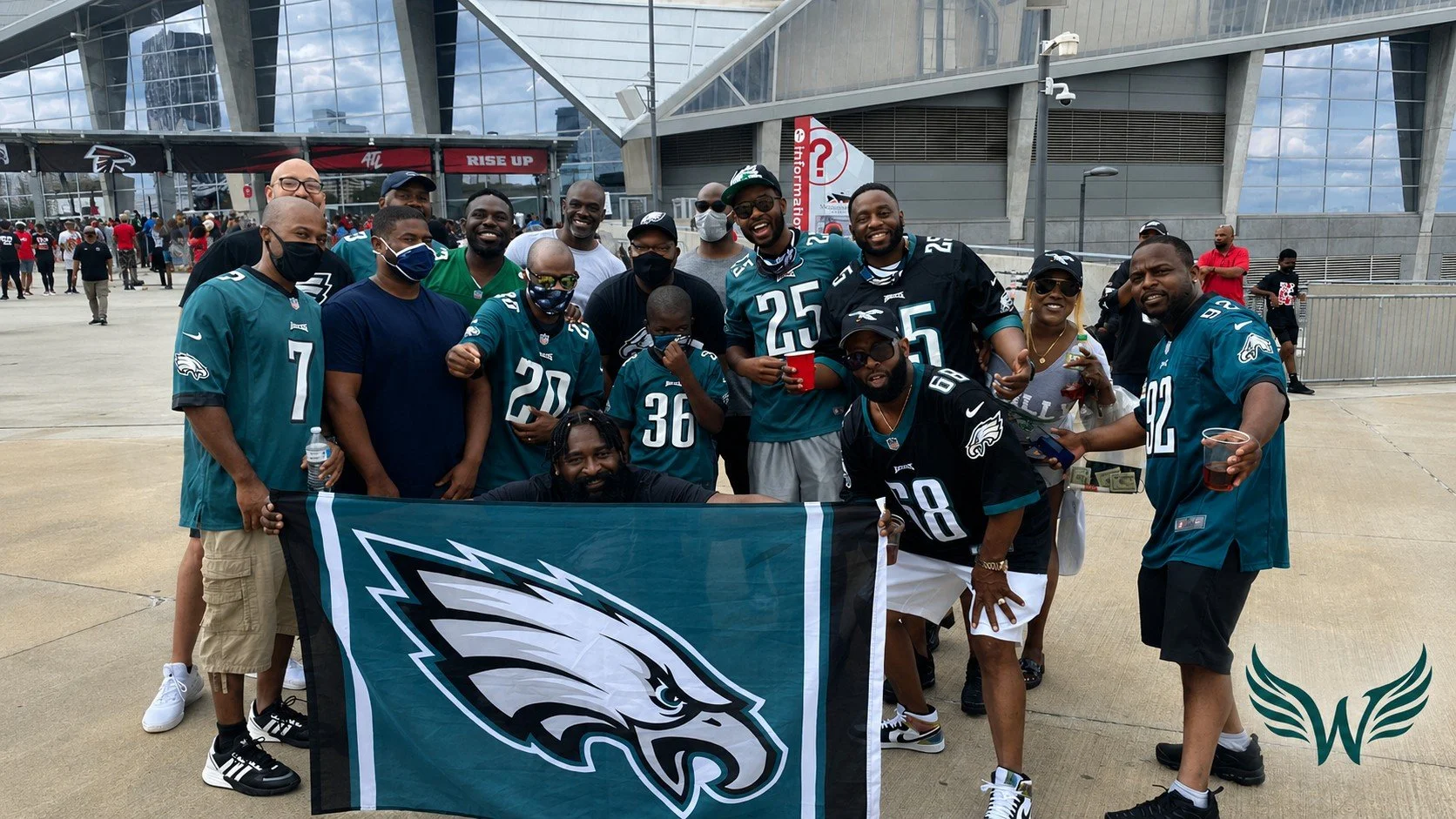 A group of football fans, dressed in Philadelphia Eagles jerseys, gathering outside a stadium, holding a Philadelphia Eagles flag, some with drinks and wearing masks, celebrating.