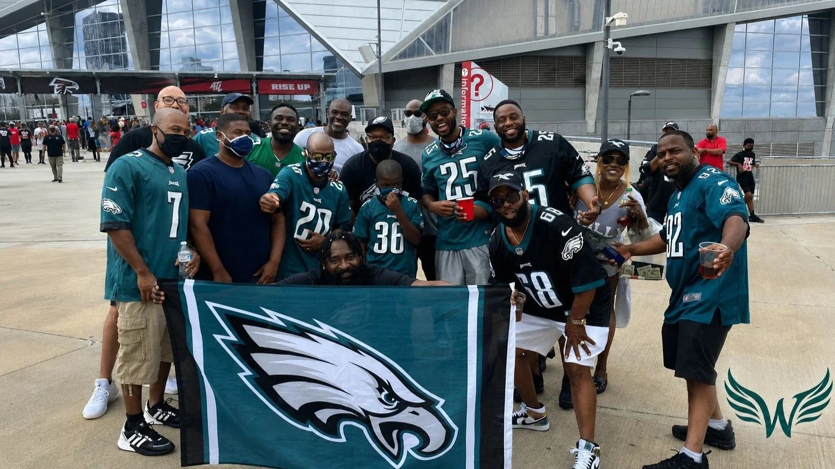 Group of people, mostly men, wearing Philadelphia Eagles football jerseys and casual clothing, posing outdoors in front of a stadium, holding a Philadelphia Eagles flag and drinks, celebrating at a football game.