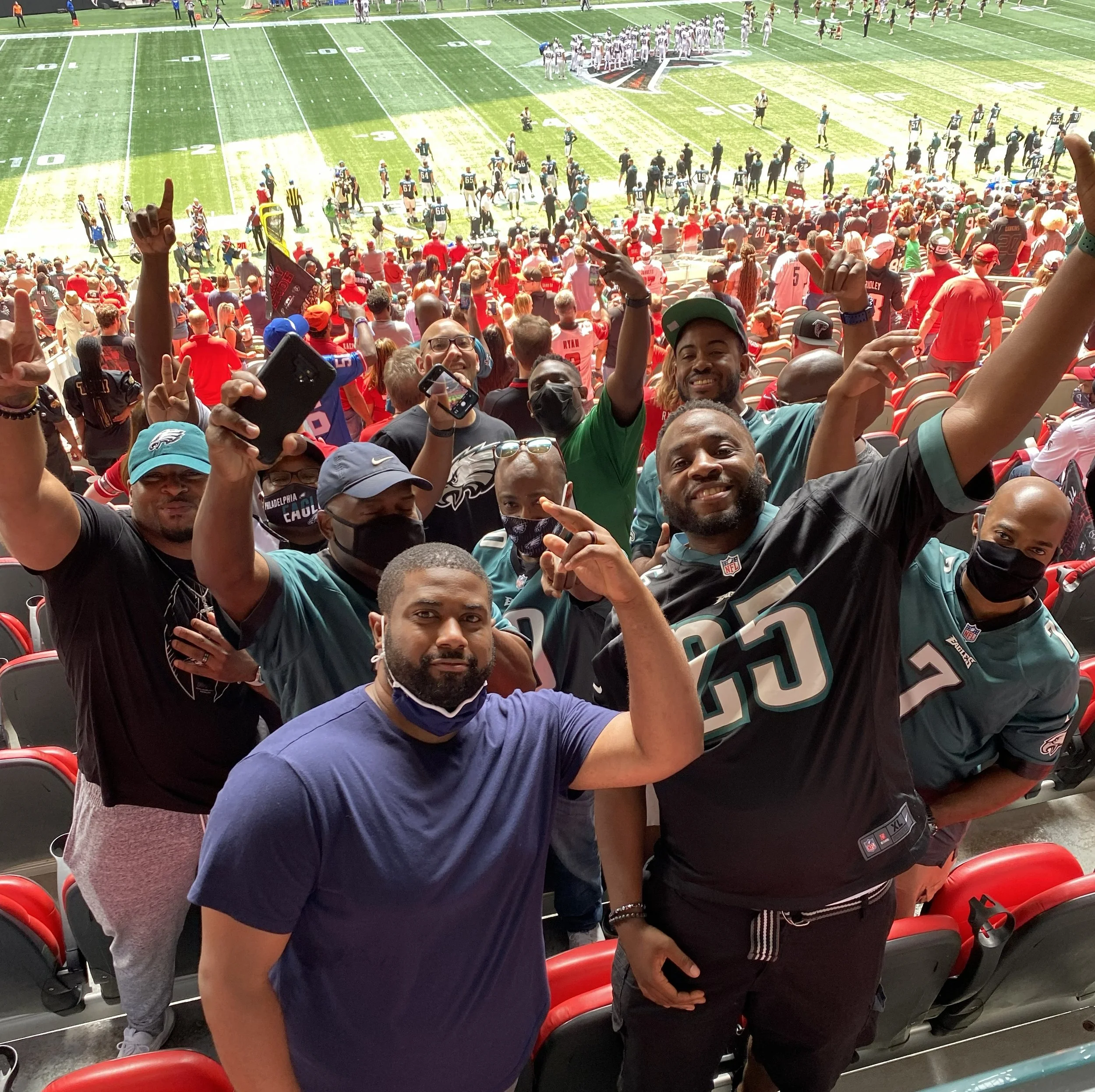 Photo of football spectators, many wearing red, black, and team jerseys, in a stadium with part of the field visible in the background. The crowd looks excited, with some taking photos and cheering.