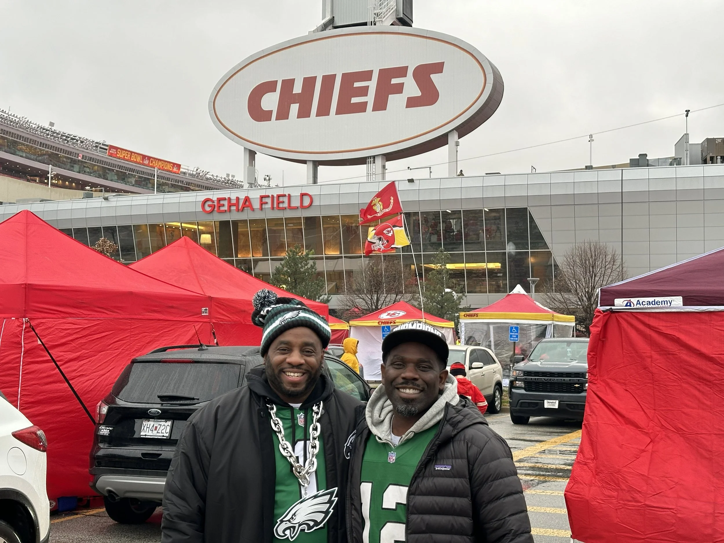 Two men smiling in a parking lot outside GEHA Field, with red tents and Philadelphia Eagles jerseys. There are multiple cars, a large Chiefs sign, and flags, indicating a sports event.